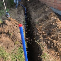 EXPERT-SALES SP Z O.O. - Wykopany rów w ziemi, z widocznym niebieskim rurociągiem z czerwoną zaślepką, pracownik w oddali i minikoparka w tle, obok budynku w trakcie budowy.