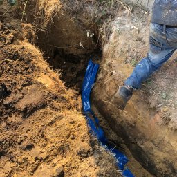 EXPERT-SALES SP Z O.O. - Wykop w ziemi wyłożony niebieską folią, widoczny fragment nogi robotnika w jeansach.