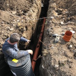 EXPERT-SALES SP Z O.O. - Instalacja rury kanalizacyjnej w wykopie; mężczyzna w roboczym ubraniu wskazuje na rurę, obok studzienka kanalizacyjna z żółtą naklejką ostrzegawczą, na ziemi narzędzia i rękawice robocze.