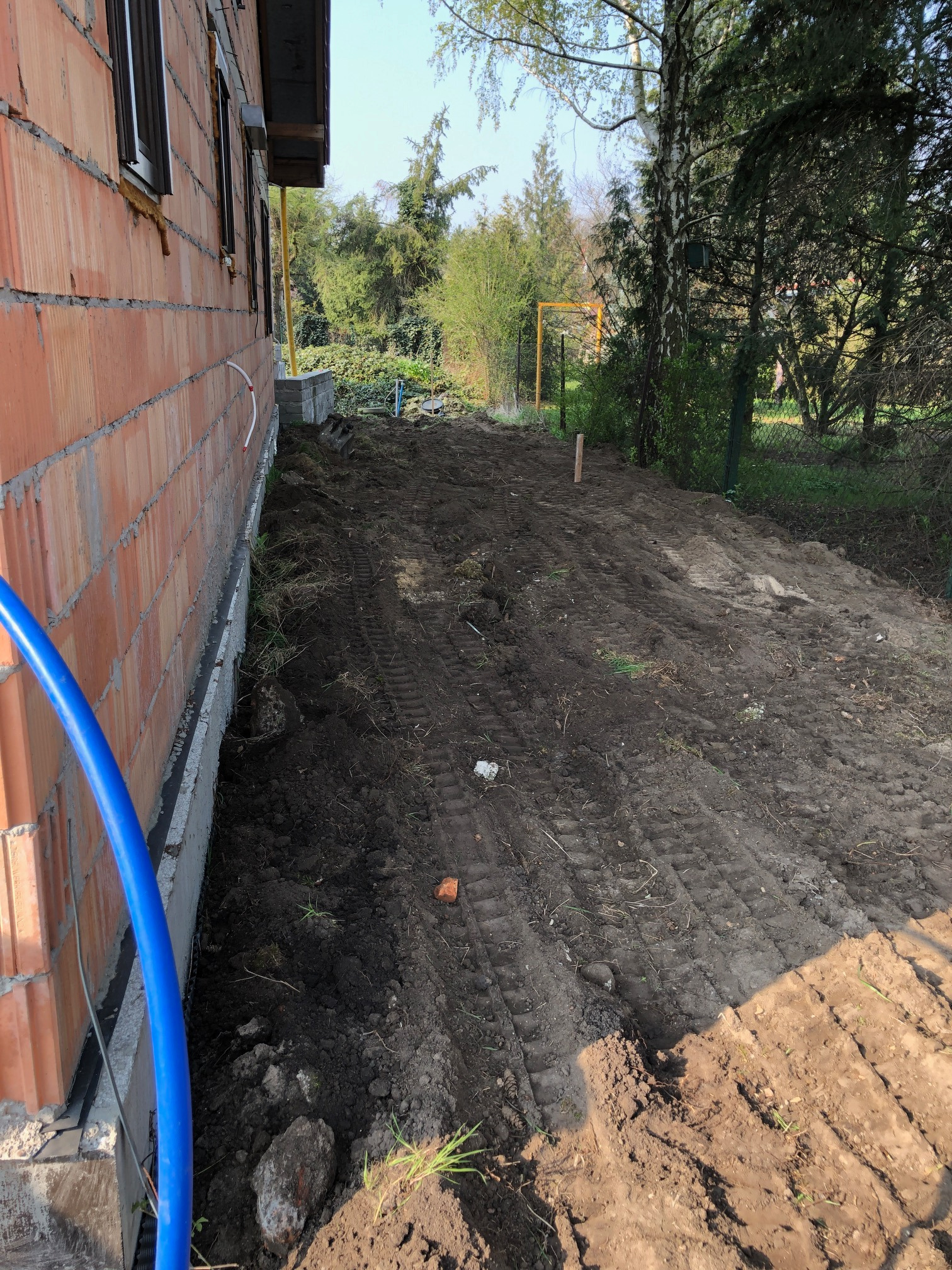 Przygotowanie terenu przy nowym budynku z cegły, widoczne ślady opon na ubitej ziemi i niebieski wąż wzdłuż fundamentów.
