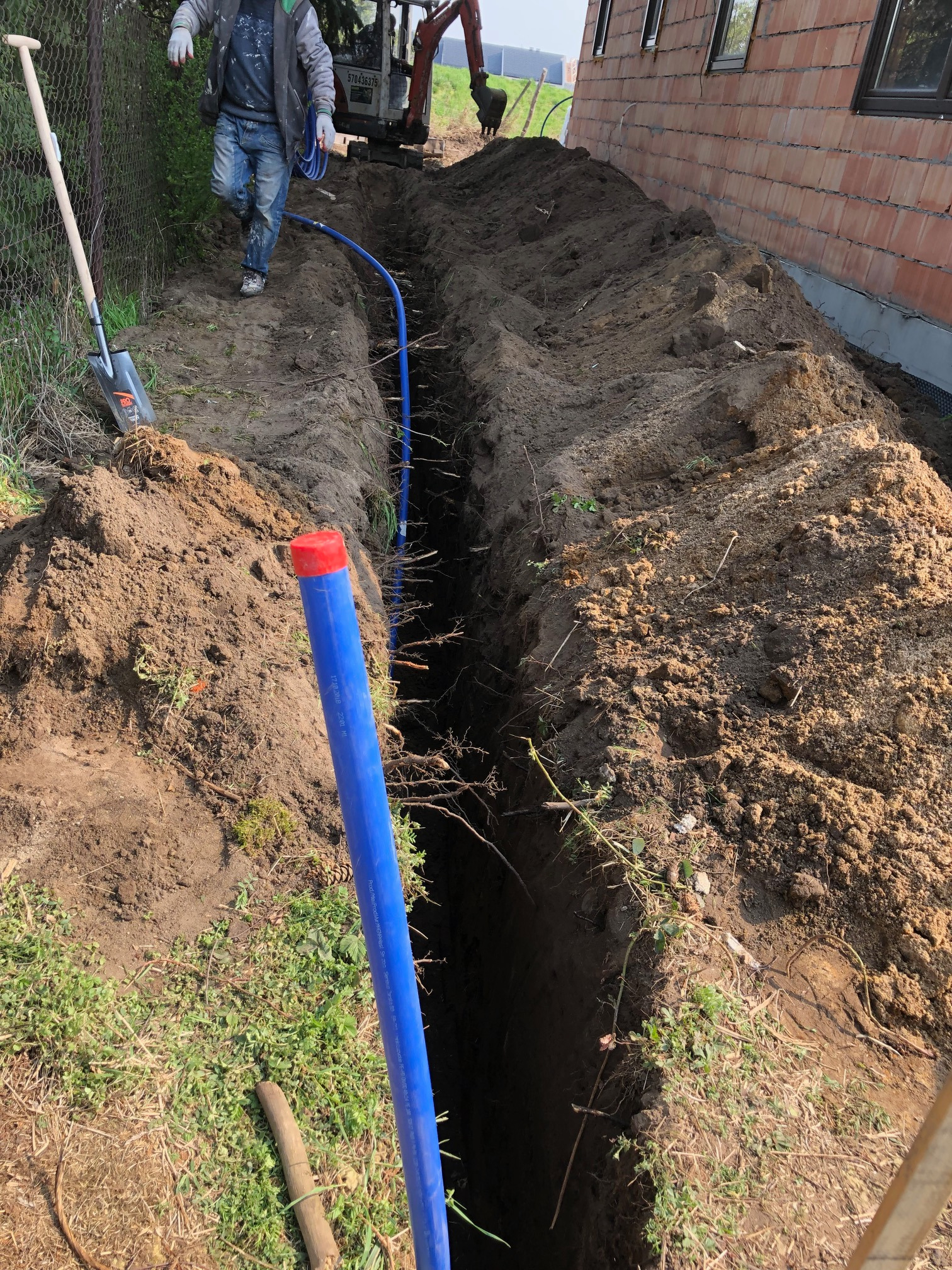 Wykopany rów w ziemi, z widocznym niebieskim rurociągiem z czerwoną zaślepką, pracownik w oddali i minikoparka w tle, obok budynku w trakcie budowy.