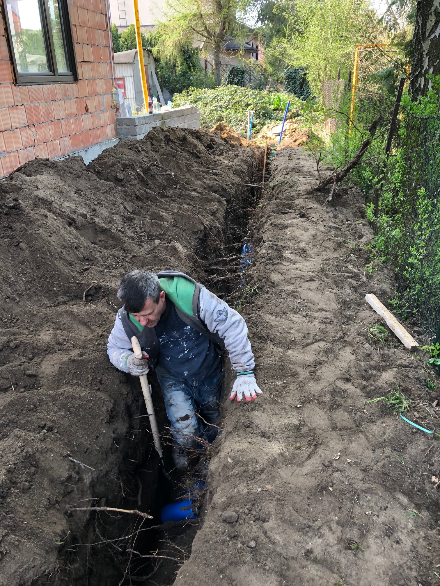 Pracownik w wykopie, wsparty na łopacie, obok rury kanalizacyjnej; widoczny fragment budynku w tle.