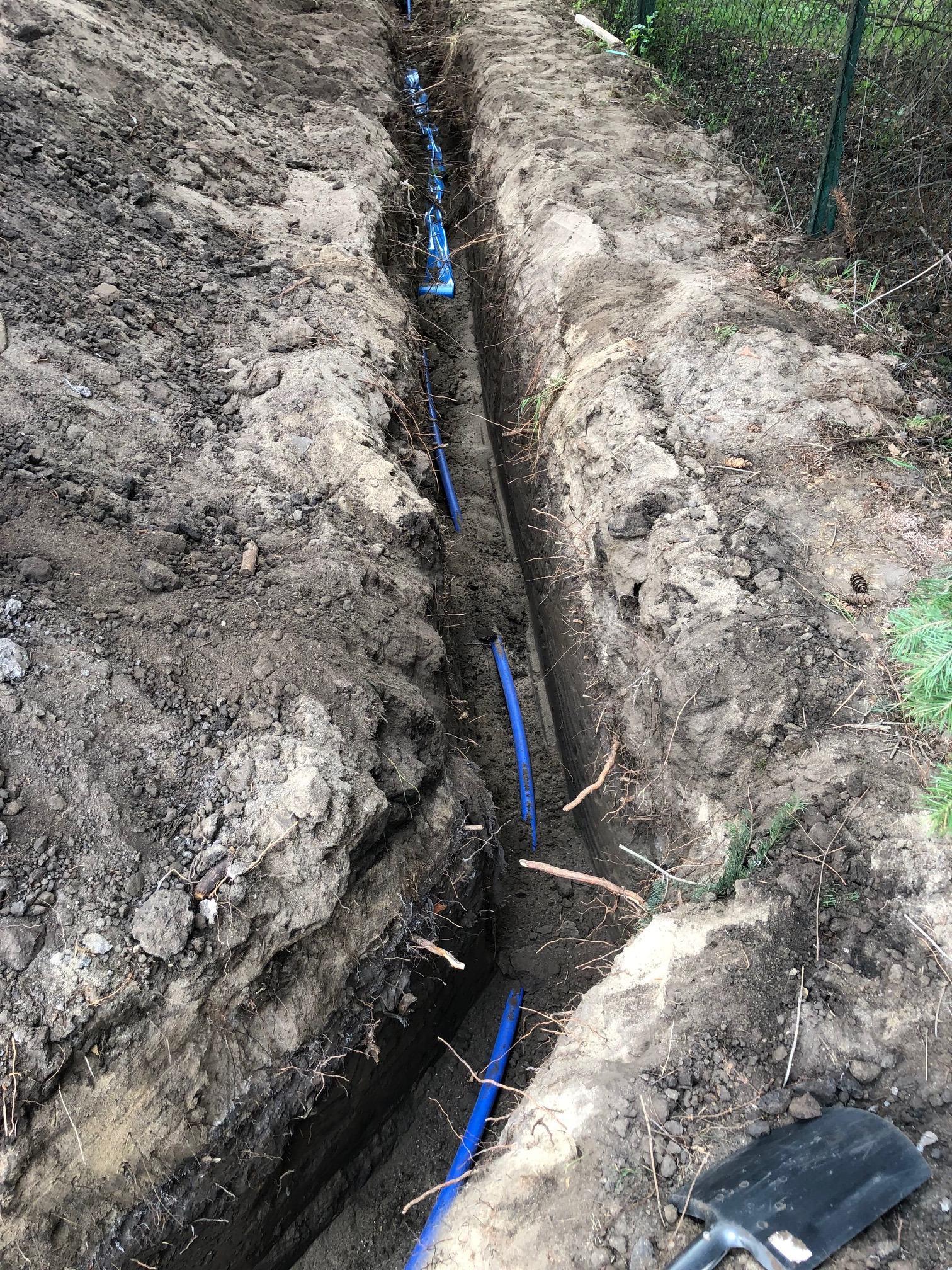 Wykopany wąski rów w ziemi z ułożonymi niebieskimi rurami, widoczna łopata na krawędzi wykopu.