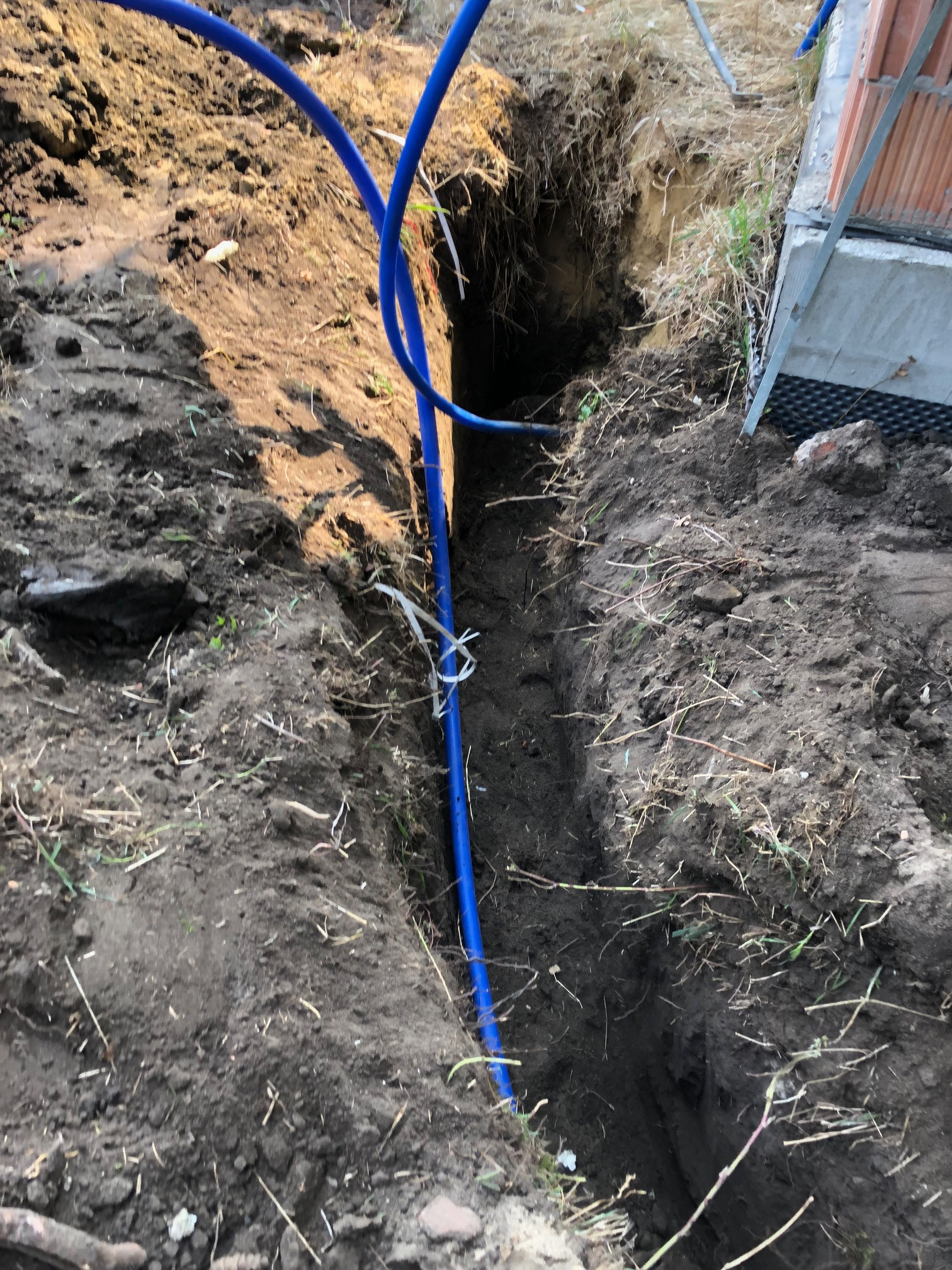 Wykopany wąski rów w ziemi z ułożoną niebieską rurą, widoczne ślady po wykopie i fragment fundamentu budynku.
