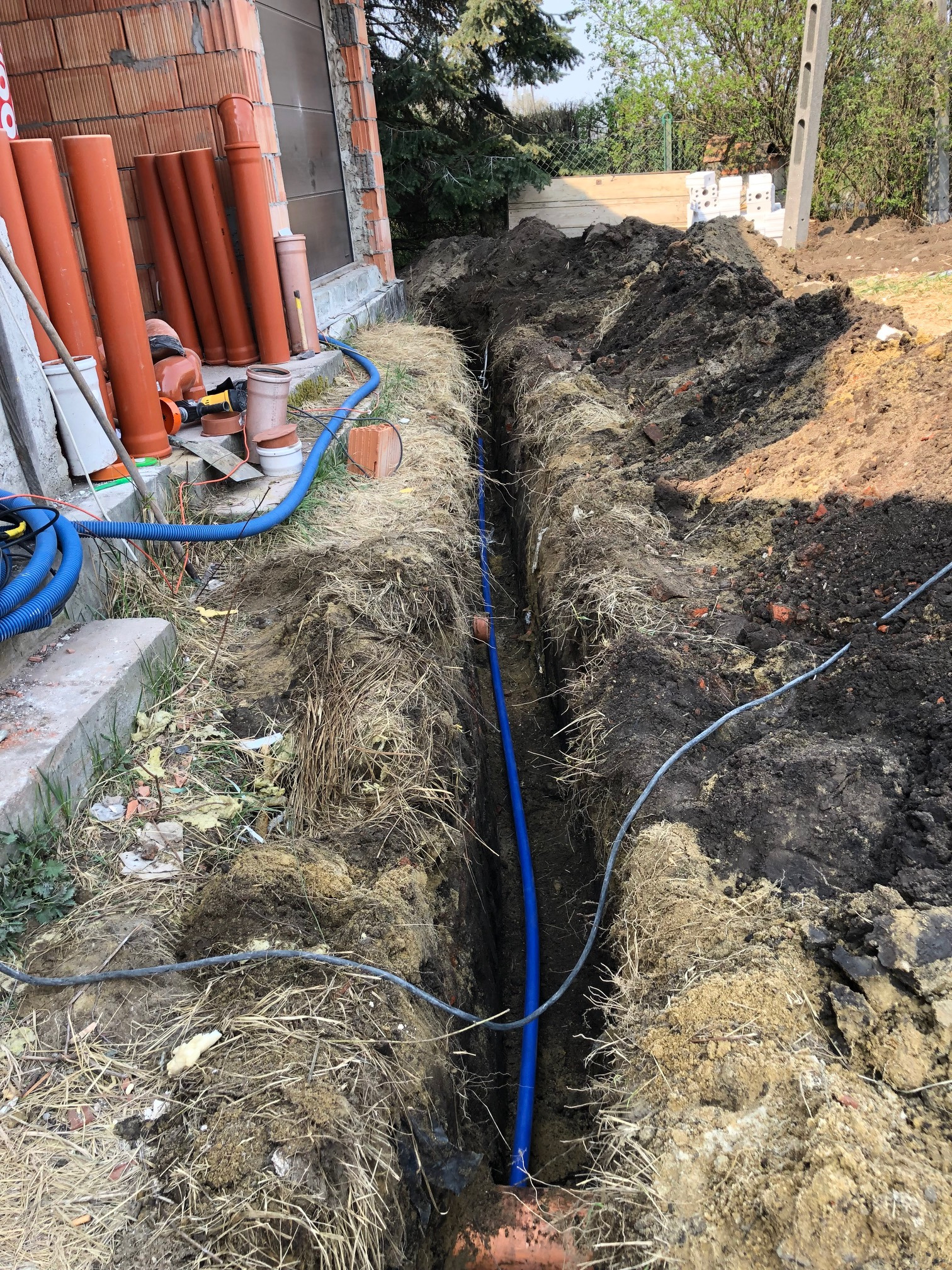 Wykopany wąski rów w ziemi z ułożoną niebieską rurą, brązowe rury kanalizacyjne i niebieski wąż na powierzchni obok budynku z czerwonej cegły.
