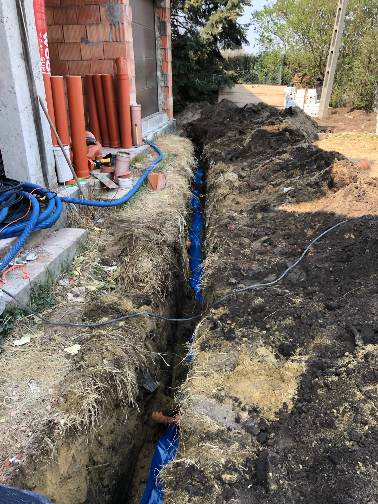 Wykopany wąski rów z niebieską folią na dnie, pomarańczowe rury na brzegu rowu, sterta ziemi obok, budynek w tle.