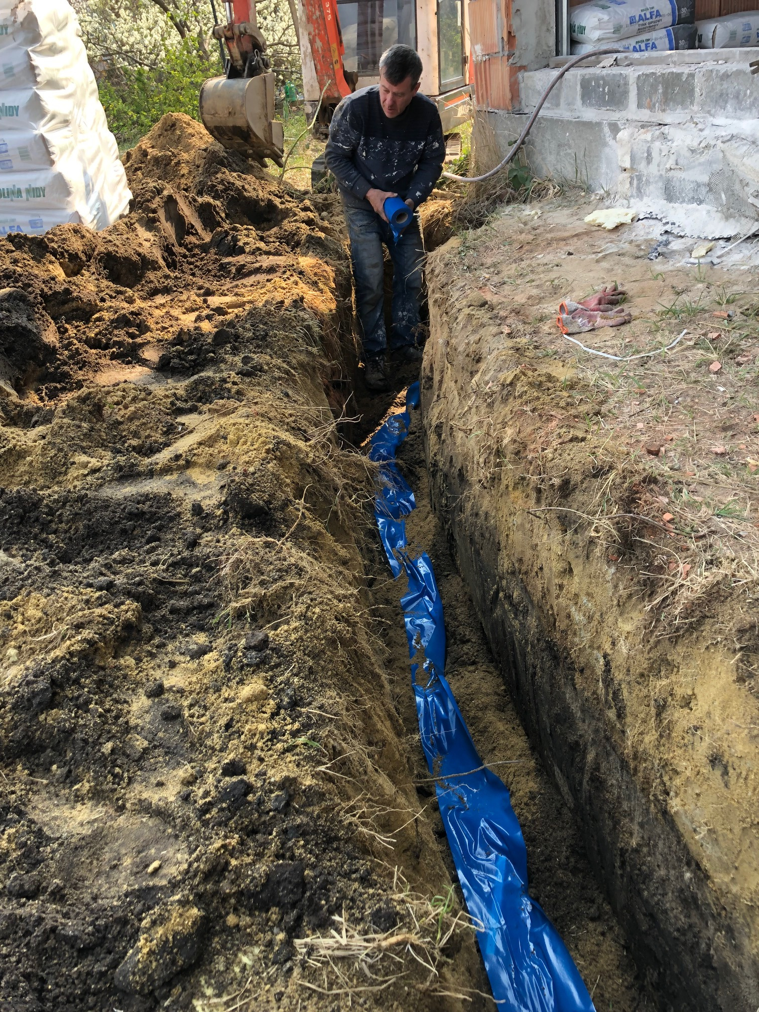 Wykopany wąski rów z niebieską folią zabezpieczającą na dnie, mężczyzna w niebieskich rękawicach stojący w wykopie, w tle mini koparka i budynek w trakcie budowy.
