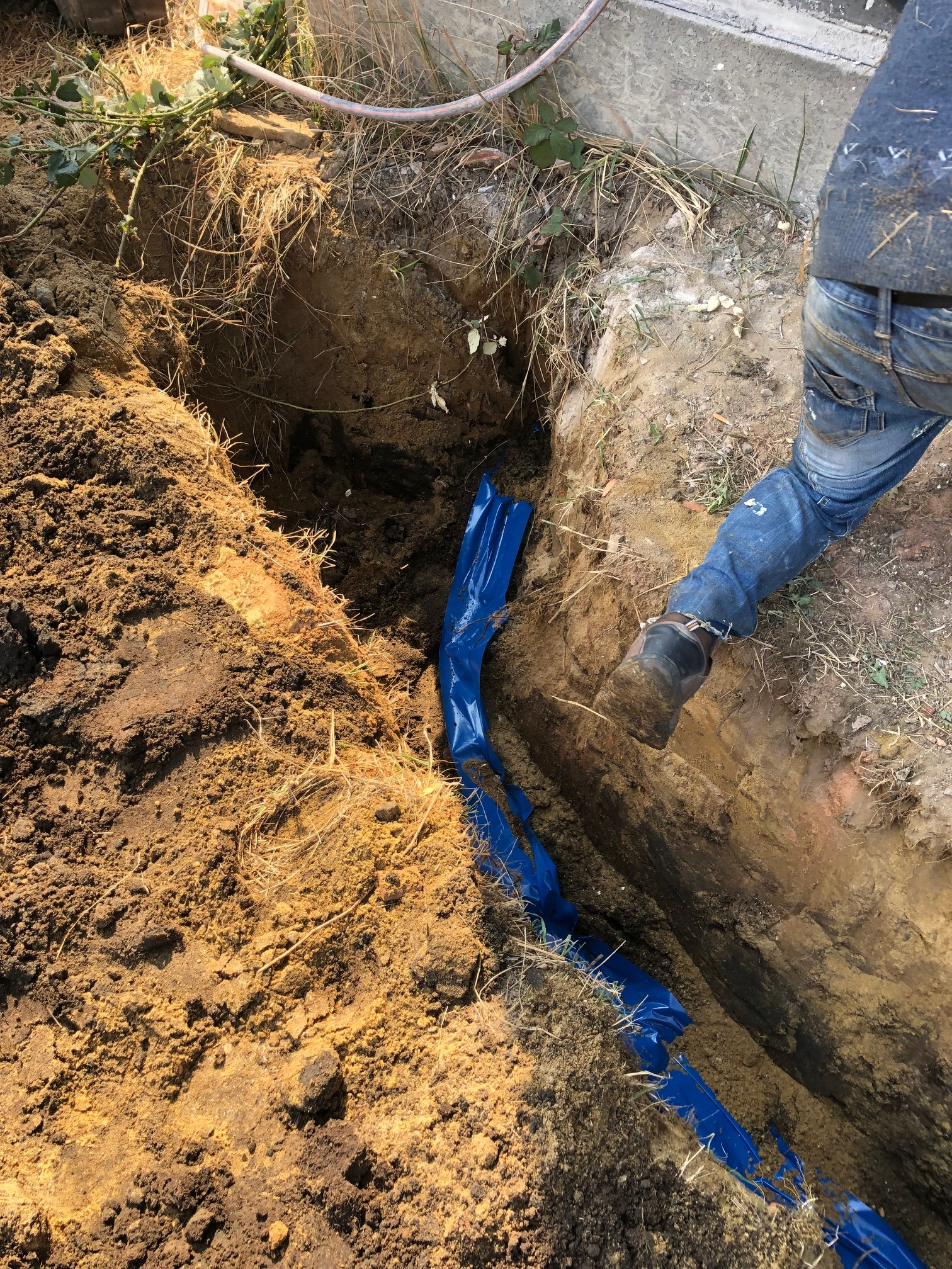 Wykop w ziemi wyłożony niebieską folią, widoczny fragment nogi robotnika w jeansach.