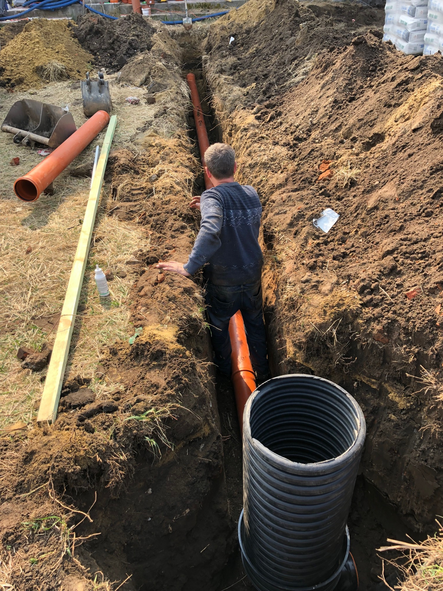Montaż pomarańczowych rur drenażowych w wykopie w Żarach. Widoczny pracownik w wykopie, rury, czarna rura karbowana, narzędzia oraz pryzma wykopanej ziemi.