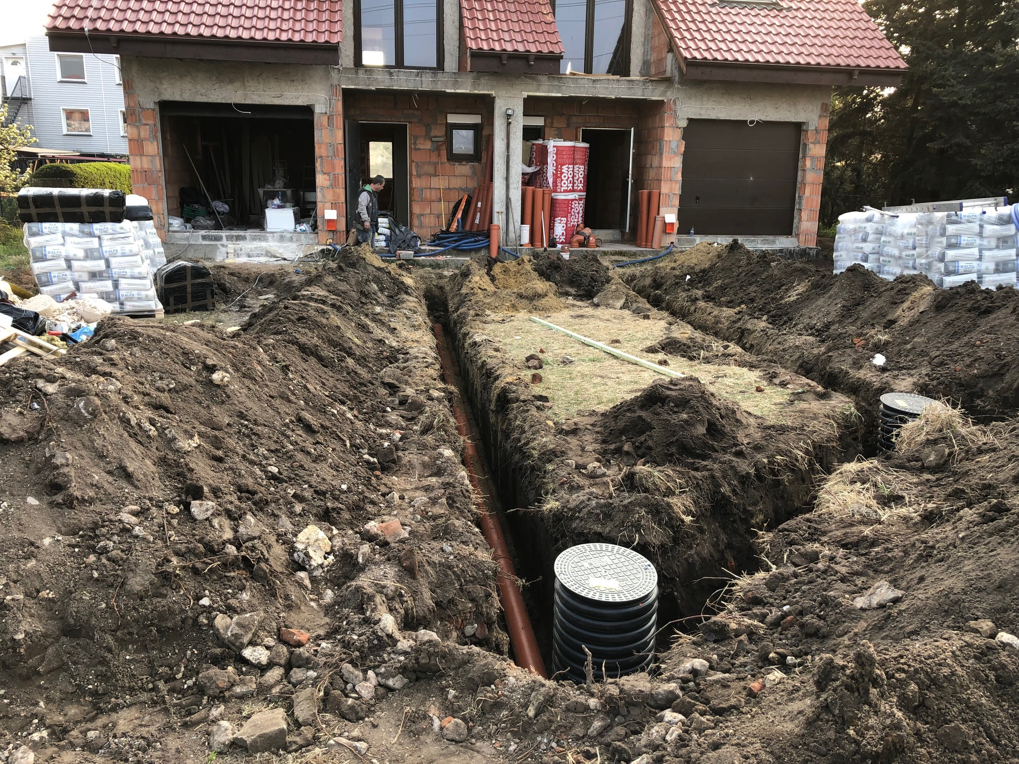 Wykopane rowy pod rury drenażowe na tle domu w trakcie budowy, widoczne worki z materiałami budowlanymi i pracownik w oddali.