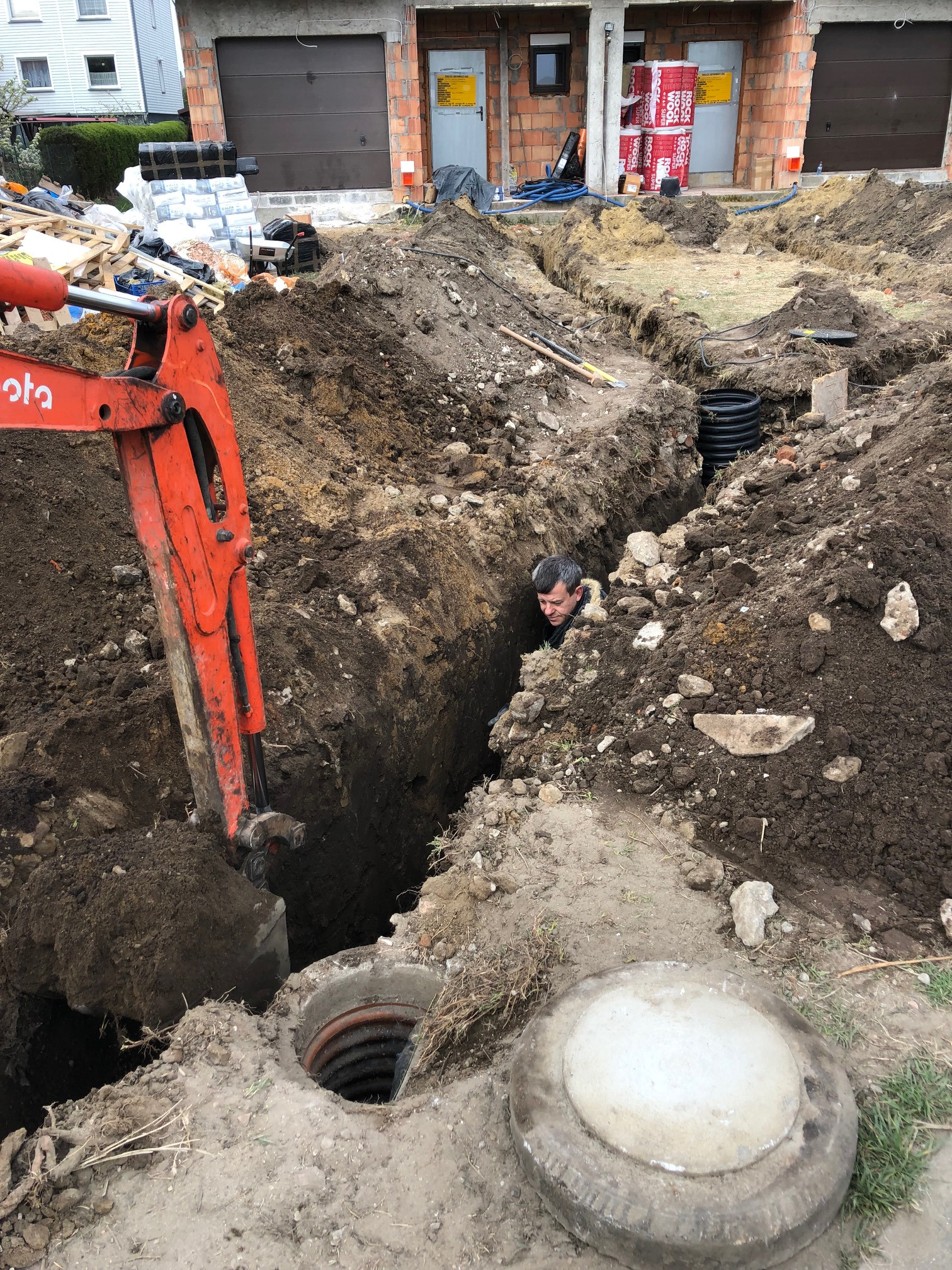 Wykop pod szambo w trakcie realizacji: pracownik w głębokim wykopie obok pomarańczowej koparki, widoczne elementy instalacji kanalizacyjnej i przygotowany otwór z betonową pokrywą.