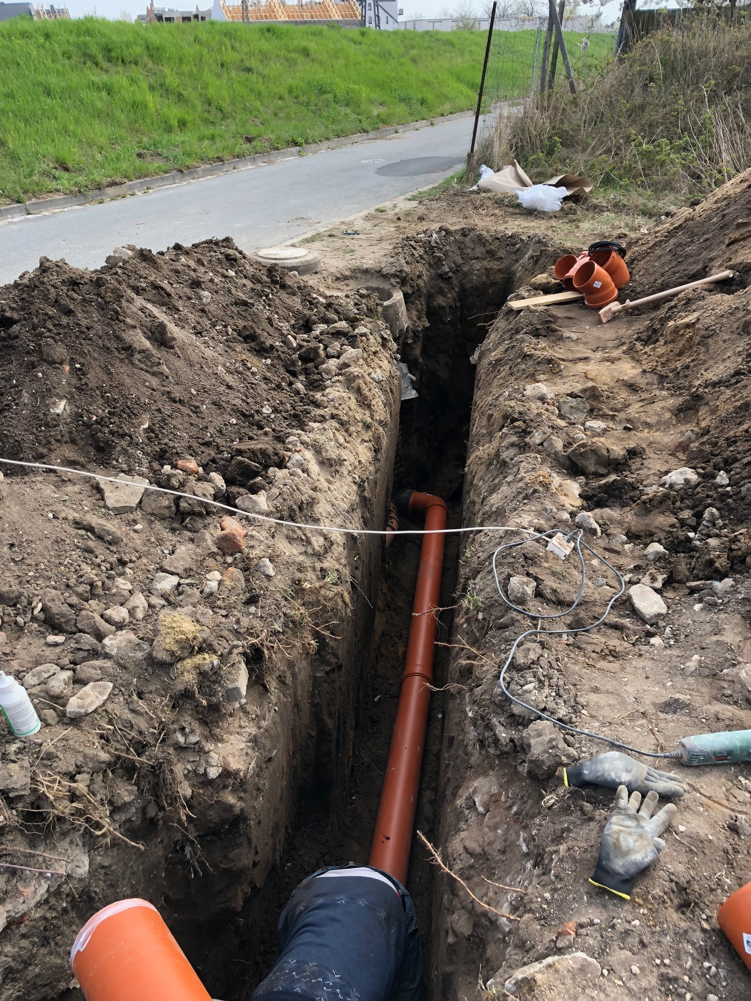 Wykop liniowy z pomarańczową rurą kanalizacyjną w środku, widoczne narzędzia i akcesoria budowlane na ziemi, w tle droga i zabudowa.