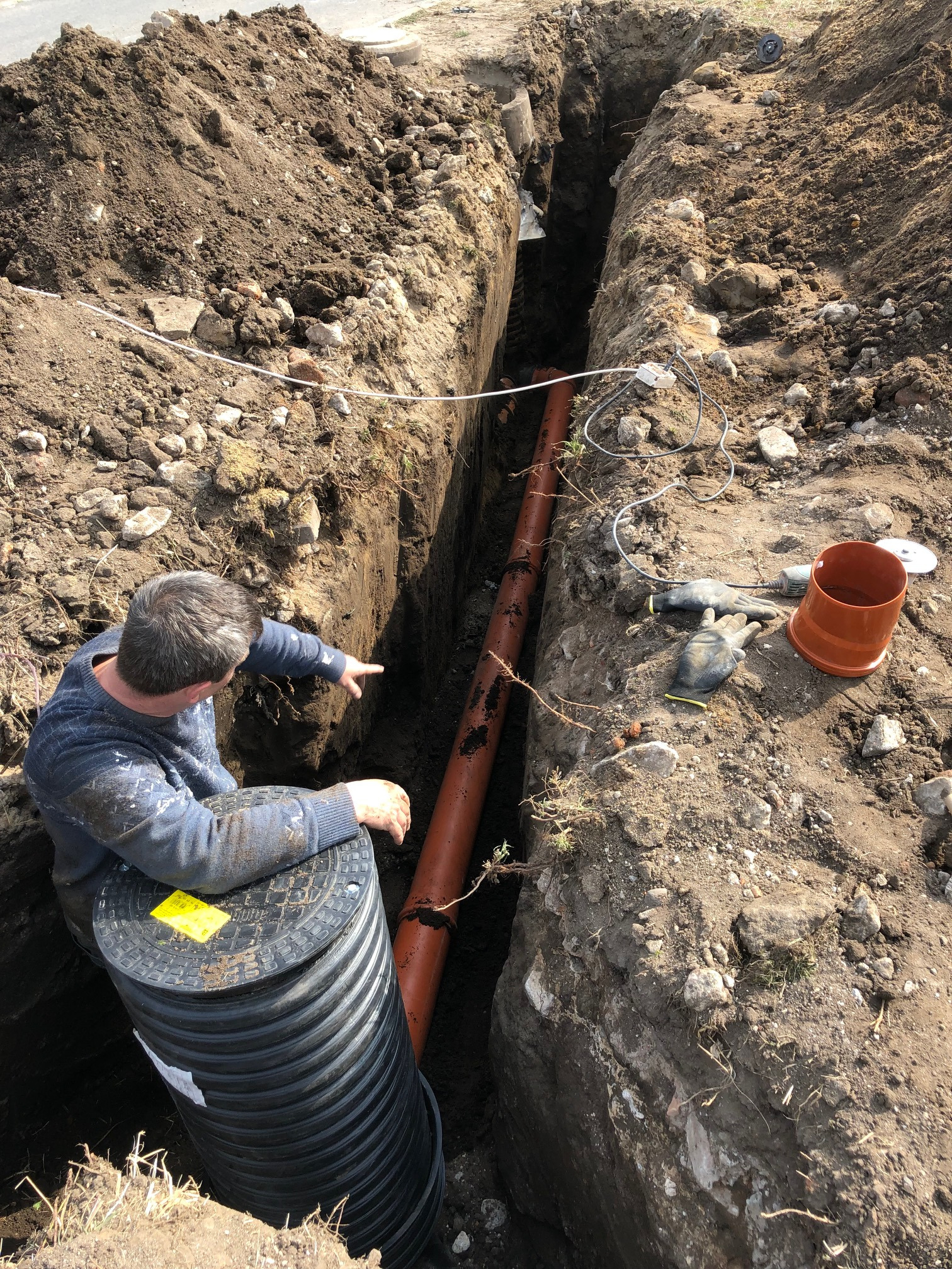 Instalacja rury kanalizacyjnej w wykopie; mężczyzna w roboczym ubraniu wskazuje na rurę, obok studzienka kanalizacyjna z żółtą naklejką ostrzegawczą, na ziemi narzędzia i rękawice robocze.