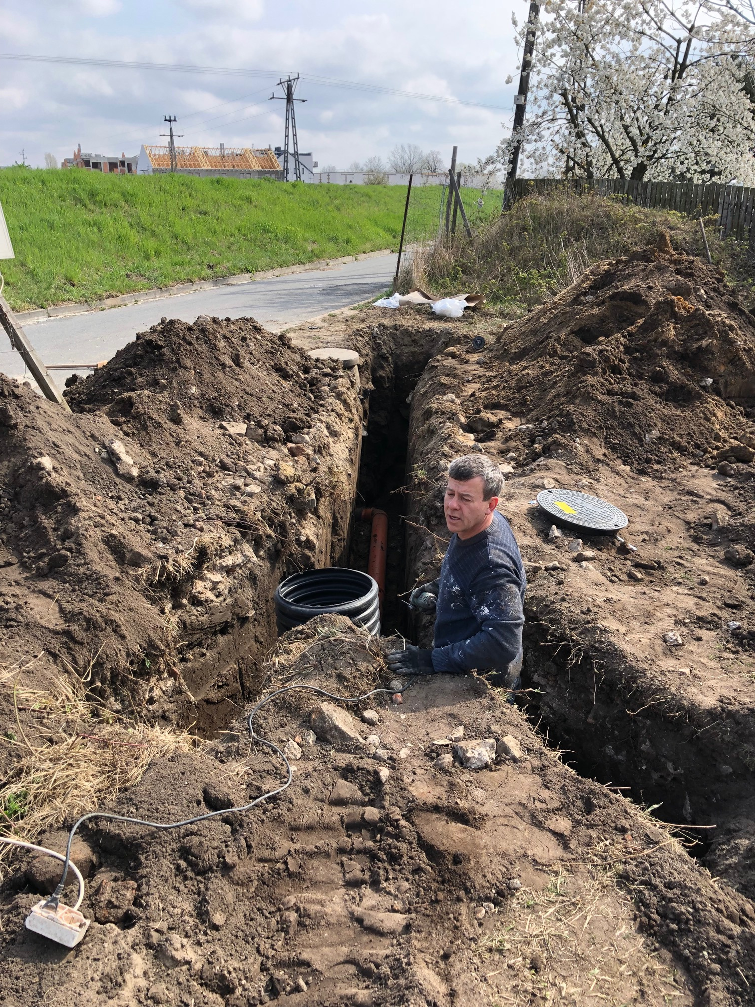 Mężczyzna w roboczym ubraniu stoi w wykopie z widocznymi rurami i czarną karbowaną rurą, obok leży właz kanalizacyjny, w tle droga i budynki.