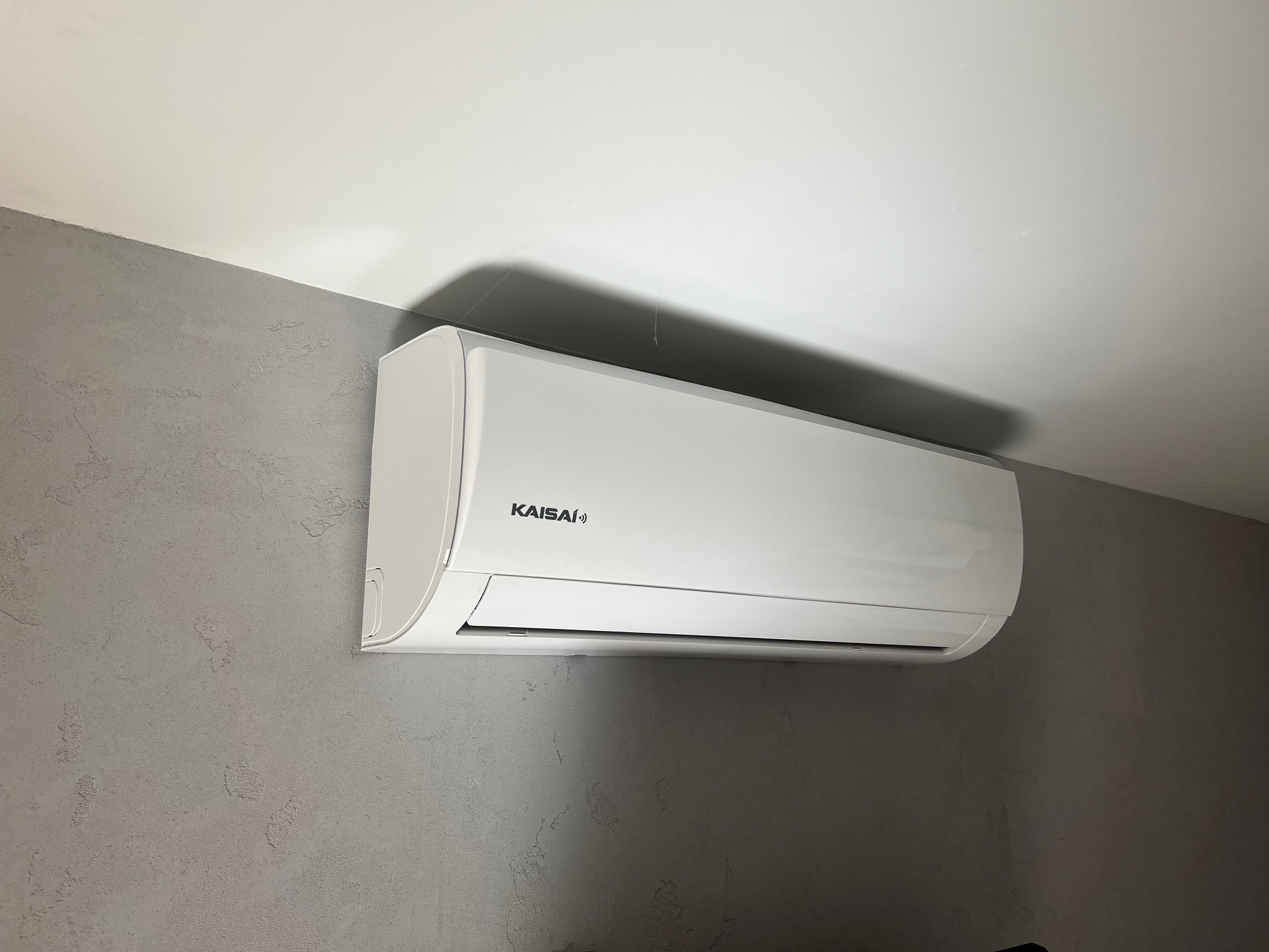 Biały klimatyzator Kaisai zamontowany na szarej ścianie o fakturze betonu, widok z dołu.