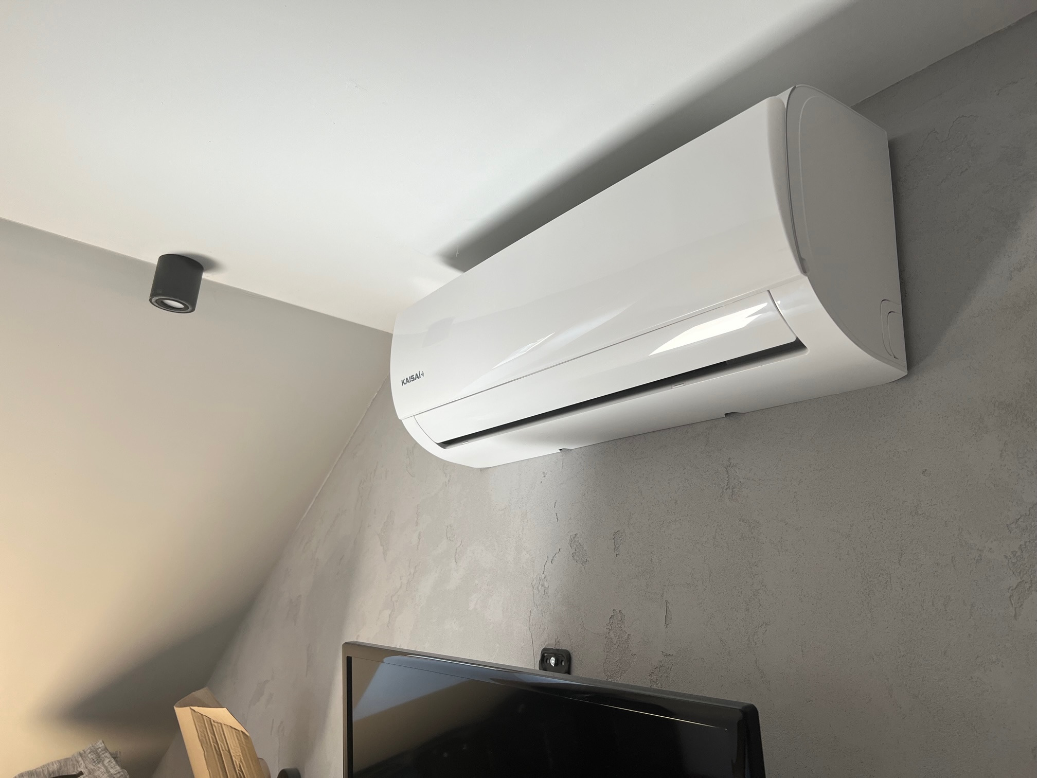 Biały klimatyzator marki Kaisai zamontowany na szarej ścianie o fakturze betonu w pomieszczeniu ze skosem, widoczny fragment telewizora i oświetlenie punktowe.