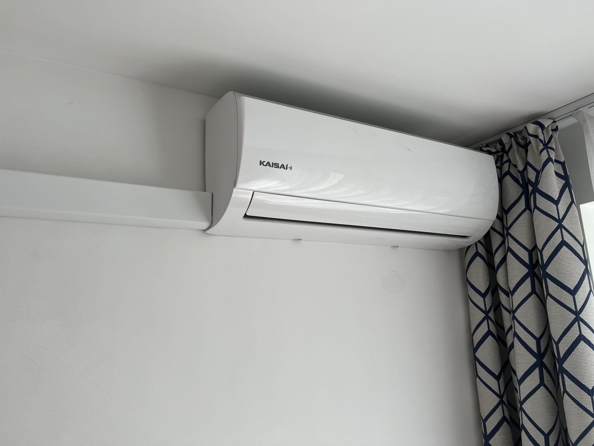Biały klimatyzator Kaisai zamontowany na białej ścianie w pomieszczeniu, widoczne zasłony z geometrycznym wzorem w odcieniach szarości i granatu.