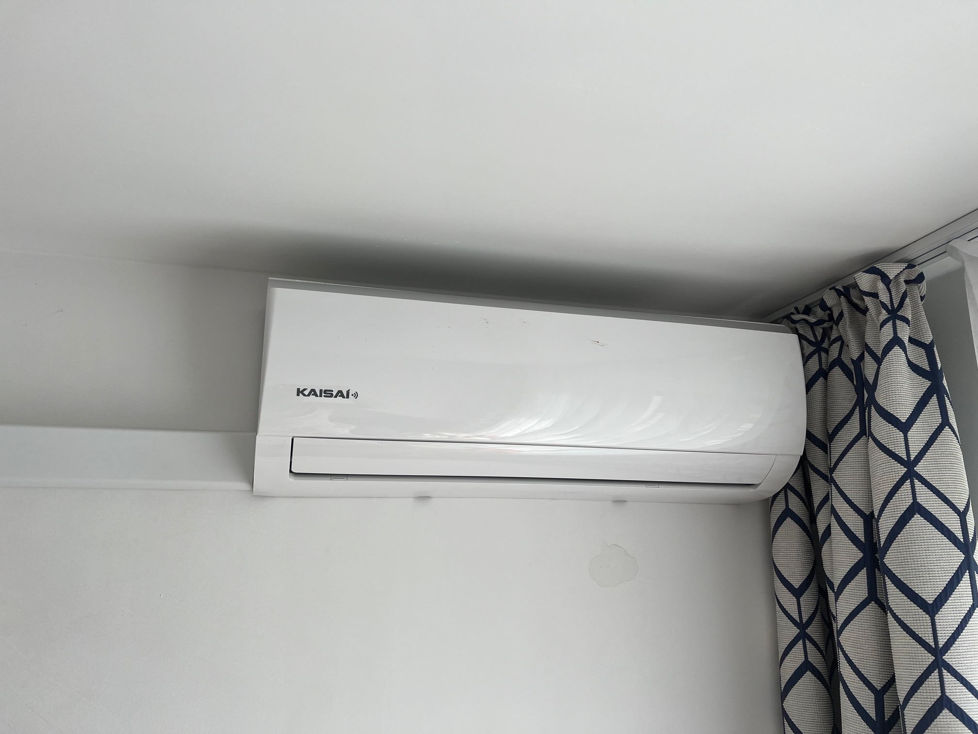 Biały klimatyzator Kaisai zamontowany na ścianie w pomieszczeniu z zasłonami w geometryczne wzory.