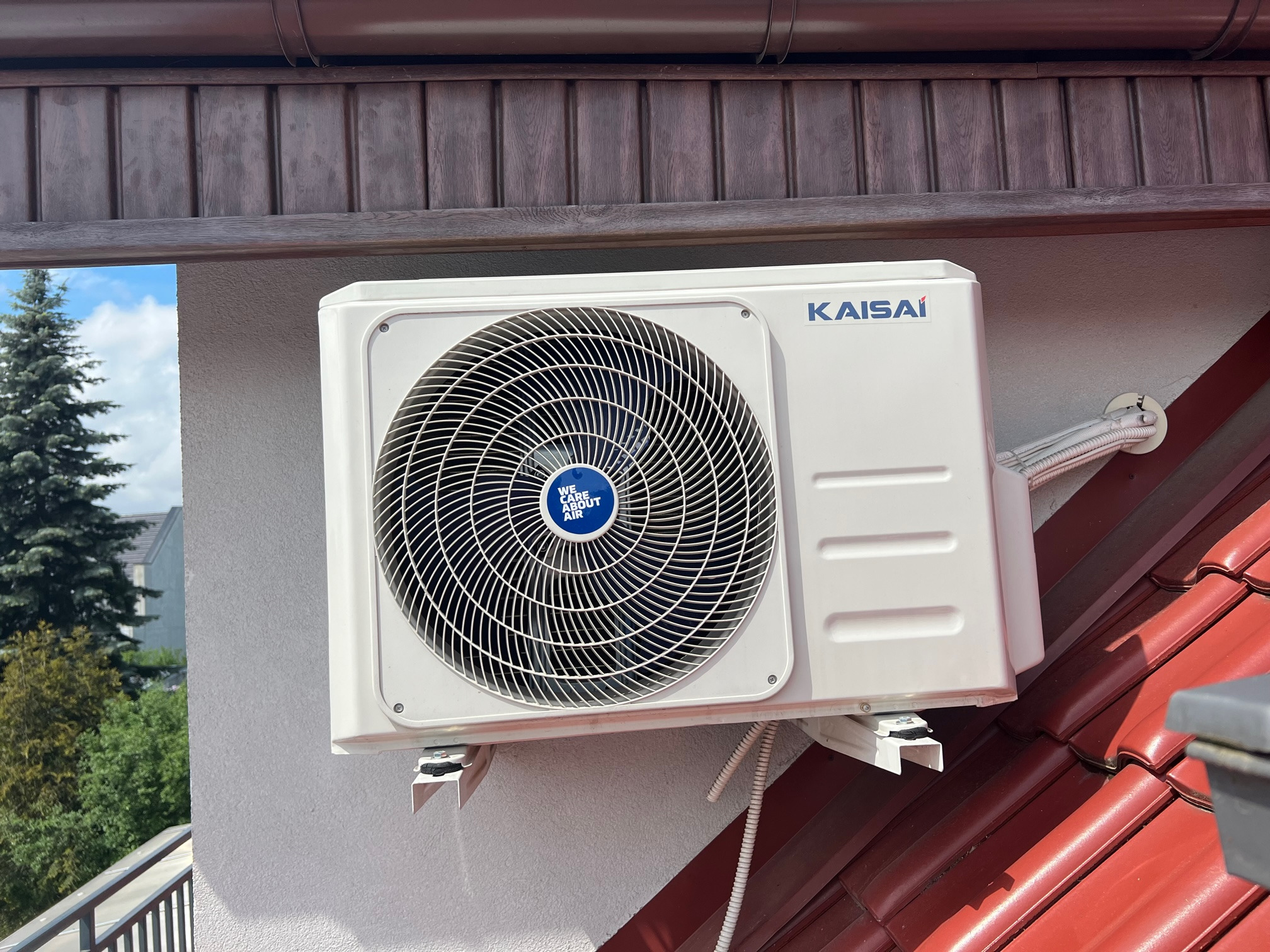Biała jednostka zewnętrzna klimatyzacji Kaisai zamontowana na ścianie domu, widoczna z bliska na tle czerwonej dachówki i drewnianej podbitki, z widocznym fragmentem nieba i drzew