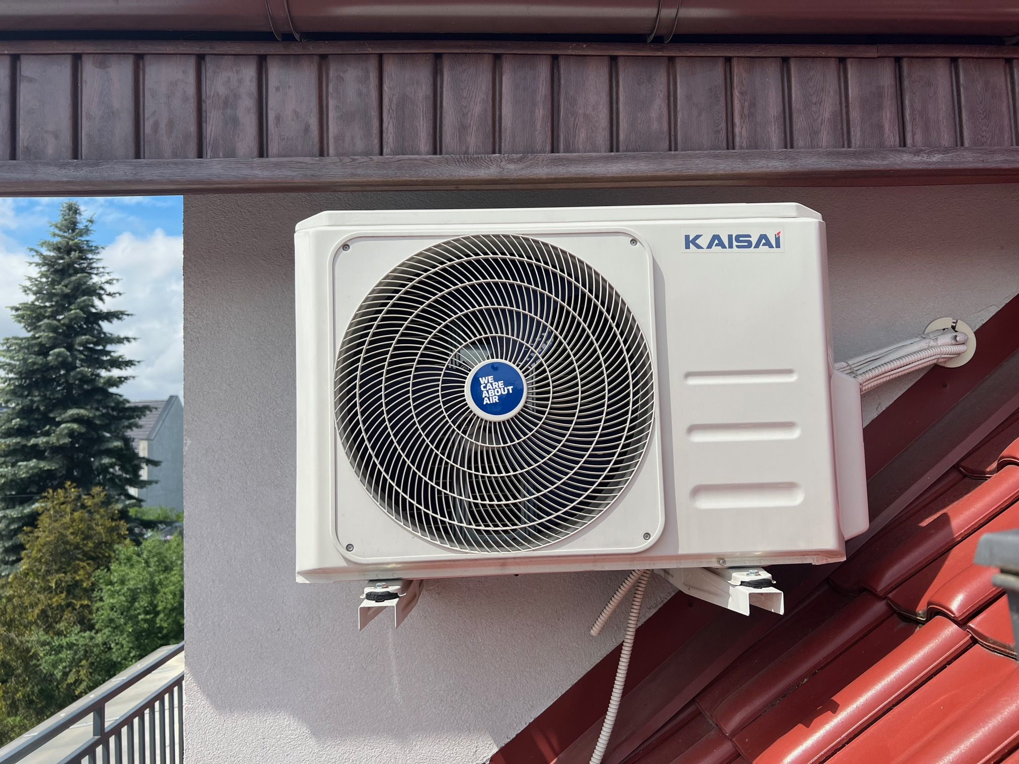 Biała jednostka zewnętrzna klimatyzacji marki Kaisai zamontowana na ścianie budynku z czerwoną dachówką, widok z boku, na tle zieleni i błękitnego nieba.