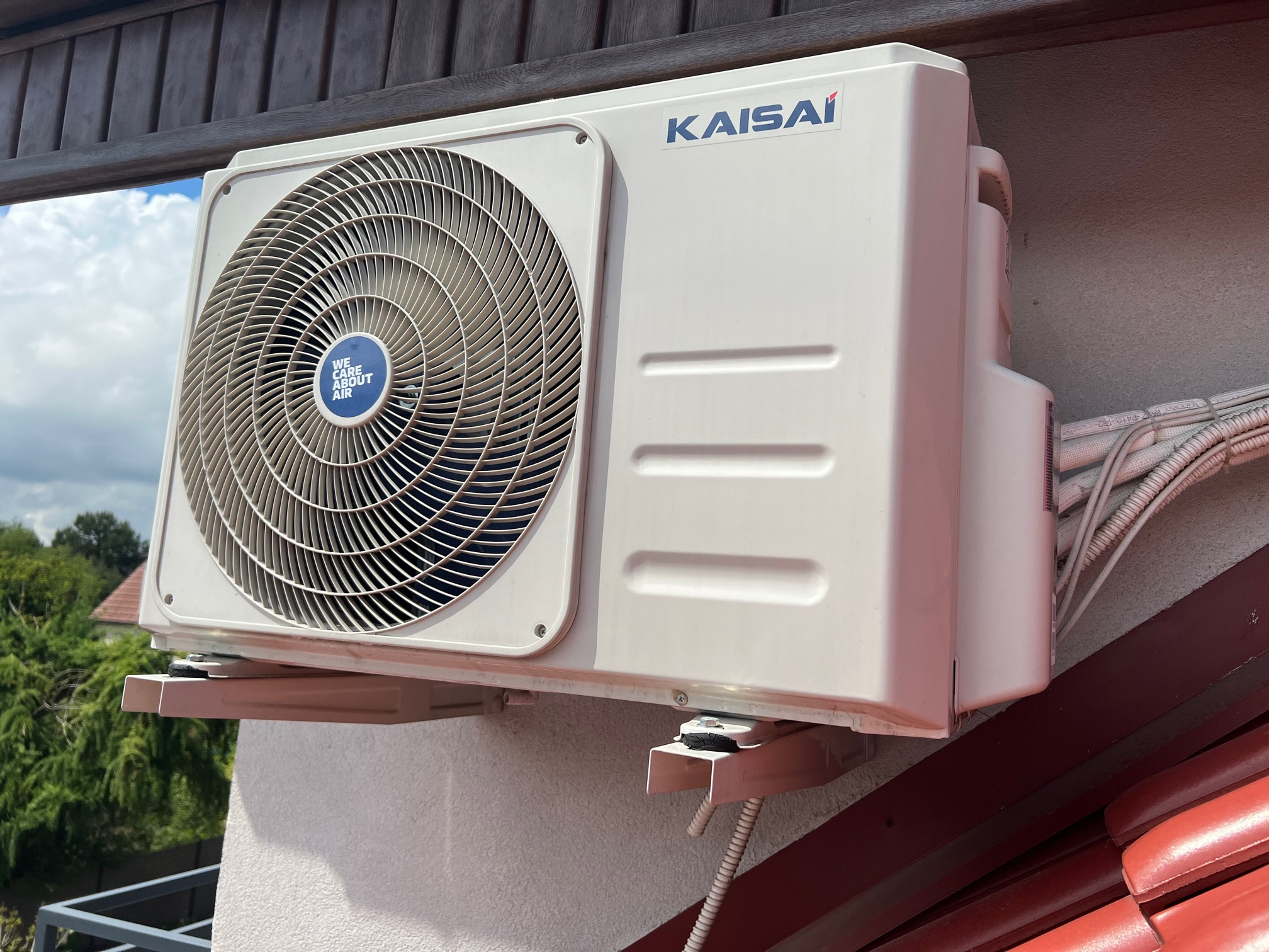 Zewnętrzna jednostka klimatyzacji Kaisai zamontowana na ścianie budynku, widoczne kable i fragment dachu z czerwonej dachówki.