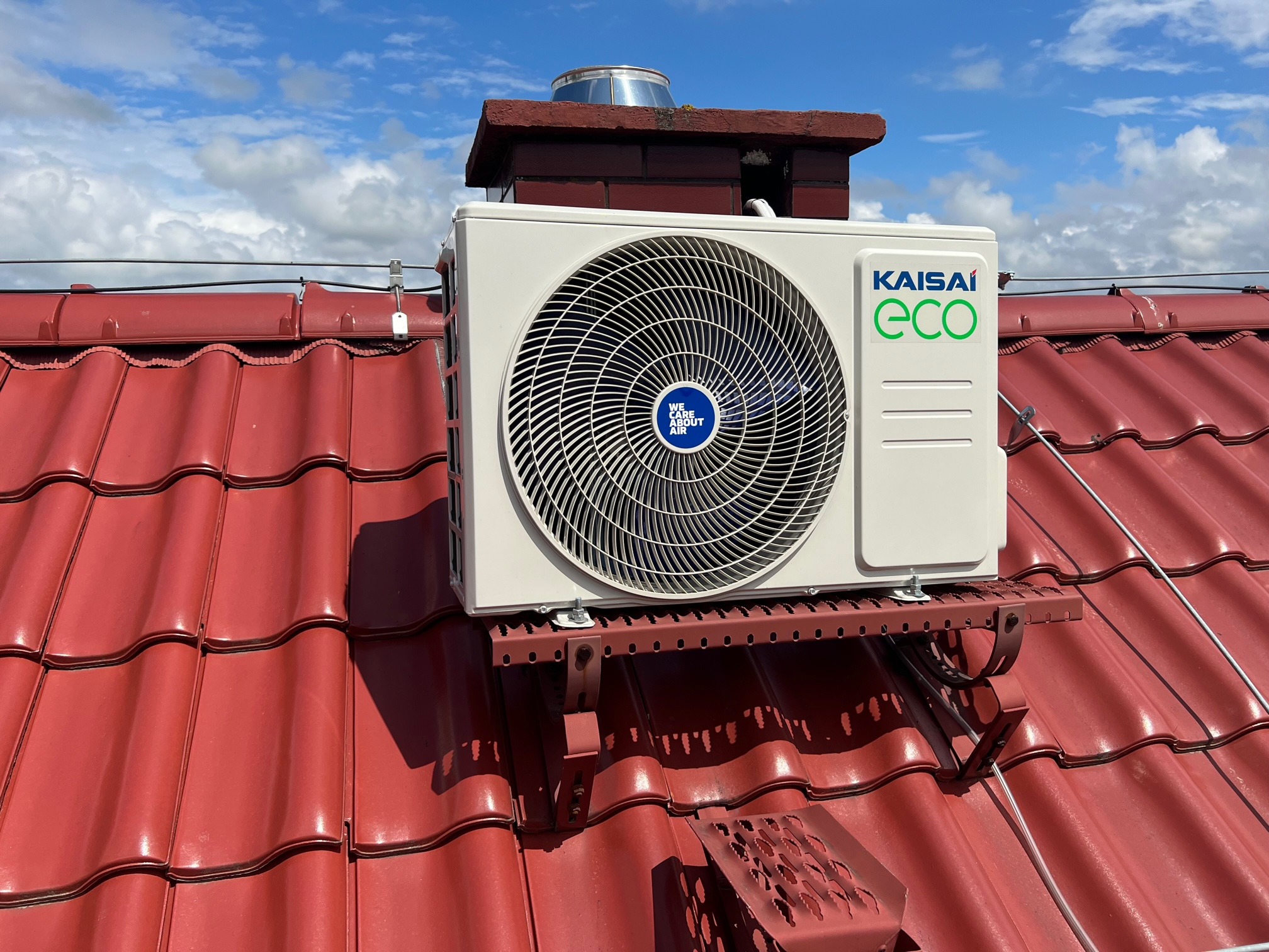 Zewnętrzna jednostka klimatyzacji Kaisai Eco zamontowana na czerwonym dachu z dachówki ceramicznej, widoczny komin i błękitne niebo z chmurami w tle.