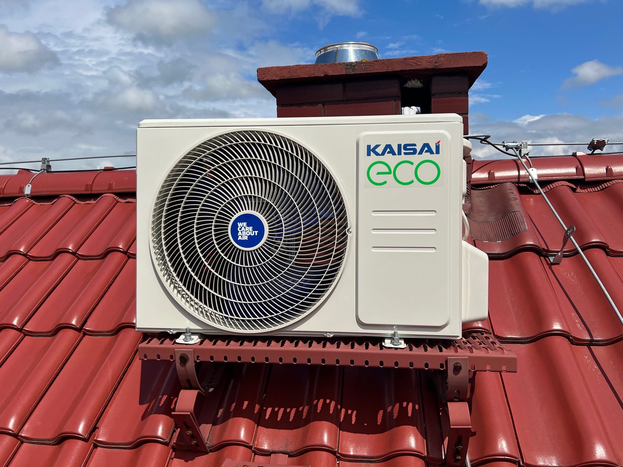 Jednostka zewnętrzna klimatyzacji Kaisai Eco zamontowana na czerwonym dachu z dachówki ceramicznej, widoczny komin i błękitne niebo z chmurami.