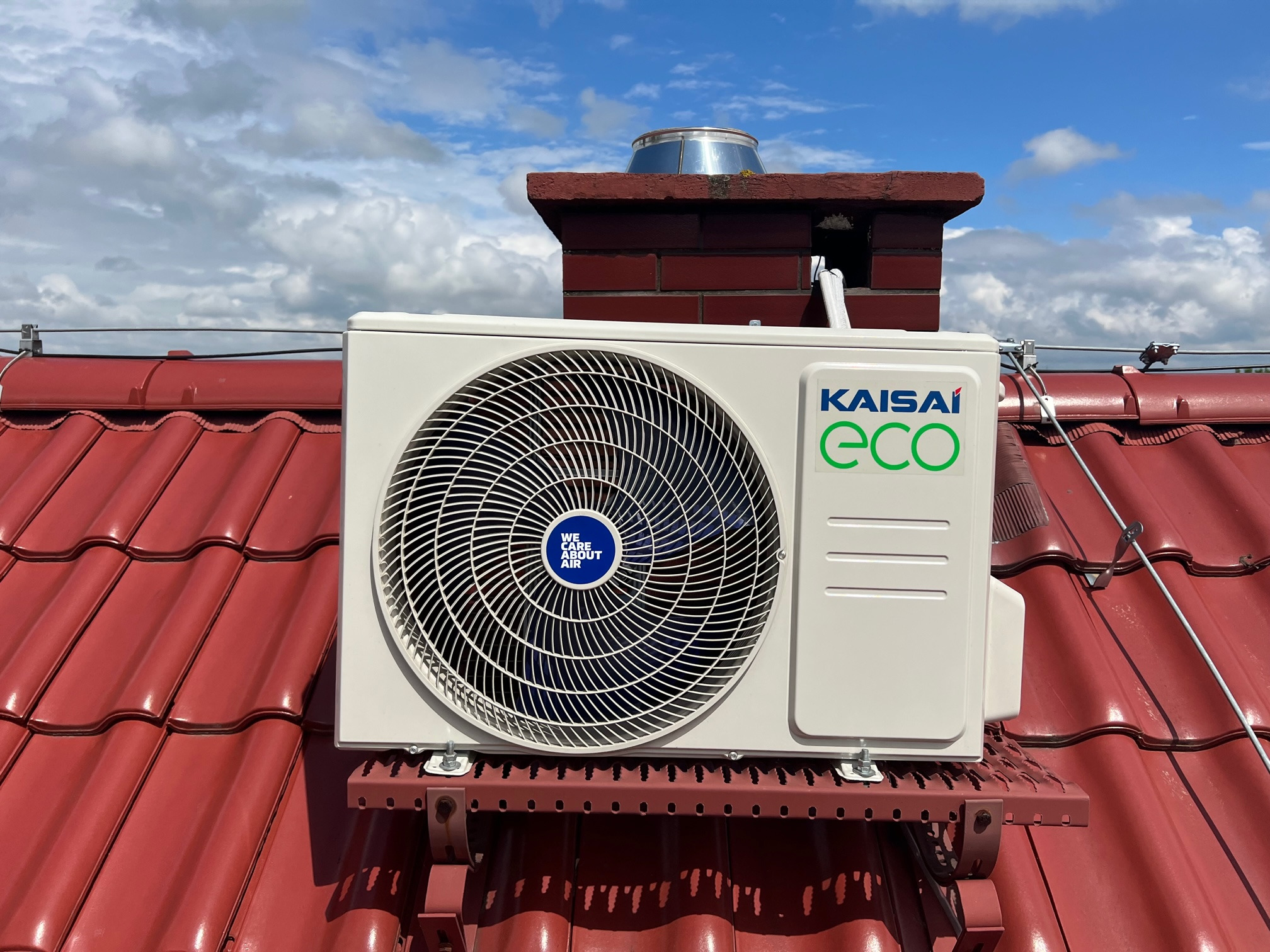 Zewnętrzna jednostka klimatyzacji Kaisai ECO zamontowana na czerwonym, spadzistym dachu z dachówki ceramicznej, widok z dołu na tle błękitnego nieba z chmurami.
