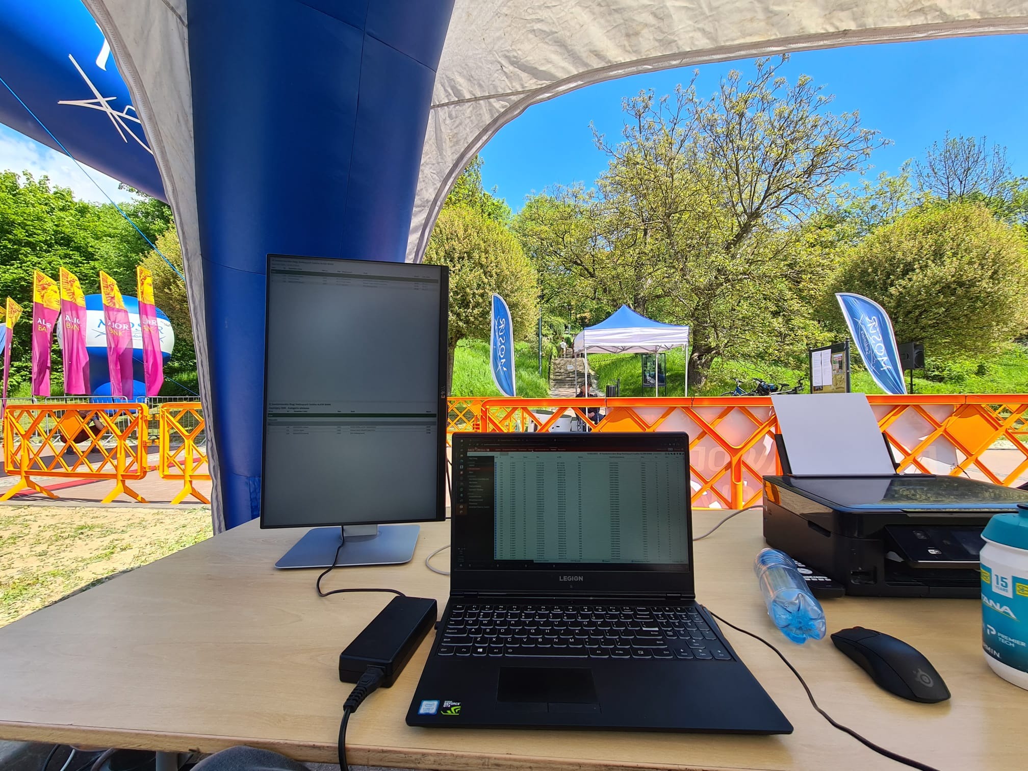 Stanowisko z laptopem, monitorem i drukarką pod namiotem podczas wydarzenia sportowego na świeżym powietrzu, widoczne wyniki na ekranach.