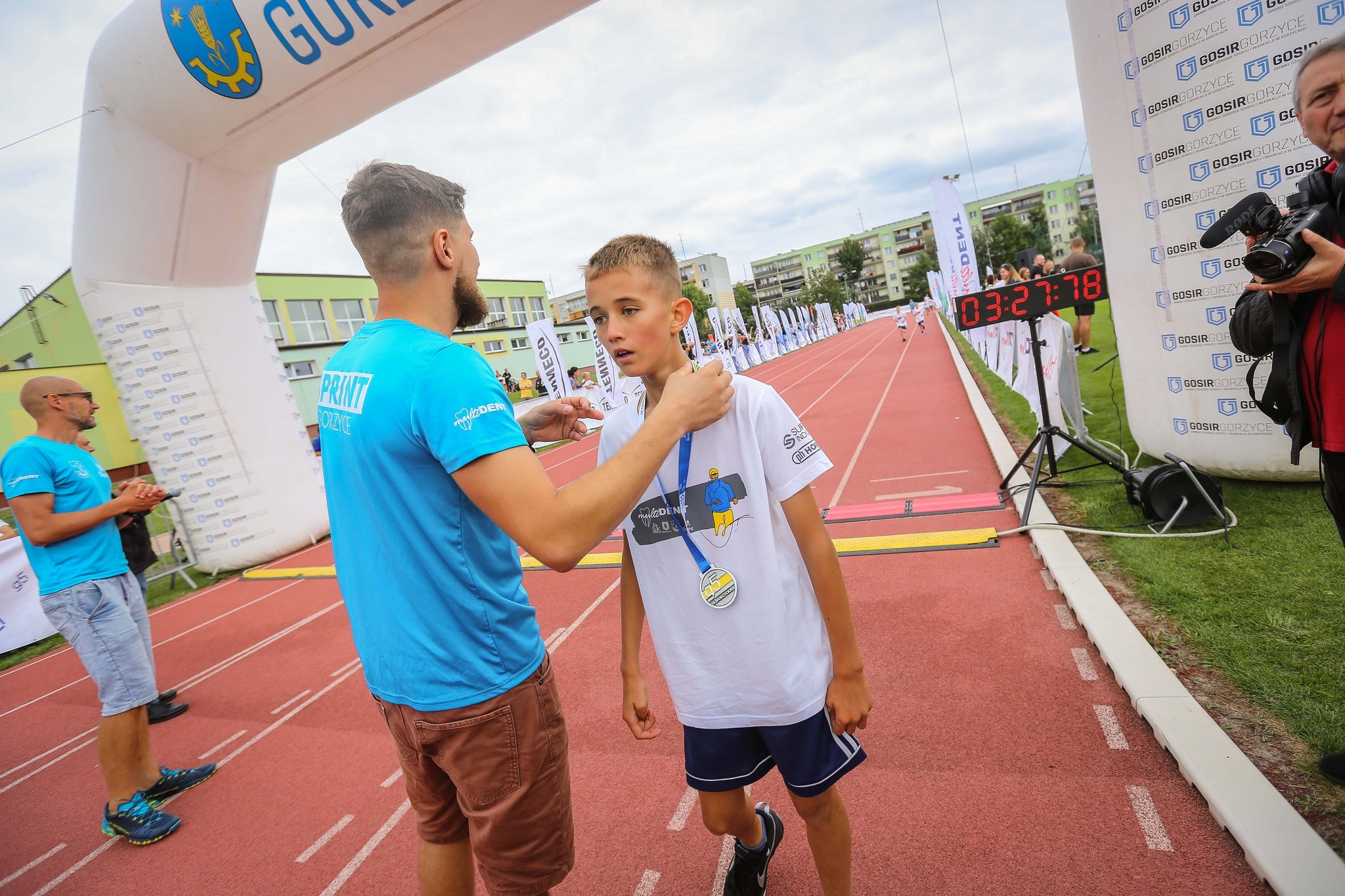 Młody biegacz z medalem na mecie, gratulowany przez trenera w niebieskiej koszulce na tle dmuchanej bramy i tablicy z czasem.
