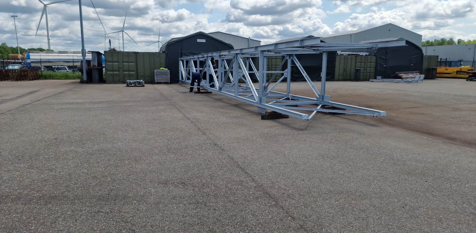 Szara stalowa konstrukcja hali na placu budowy, w tle kontenery, budynki magazynowe i turbiny wiatrowe.