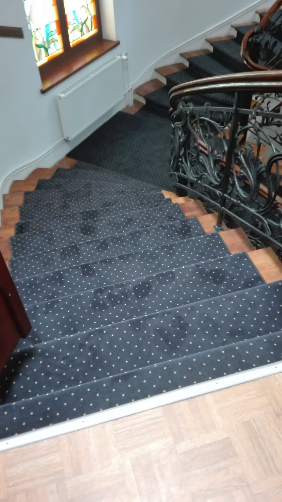 Wewnętrzne schody z ciemną wykładziną w złote kropki, drewniane stopnie i ozdobna, metalowa balustrada z roślinnymi motywami, widok z góry.