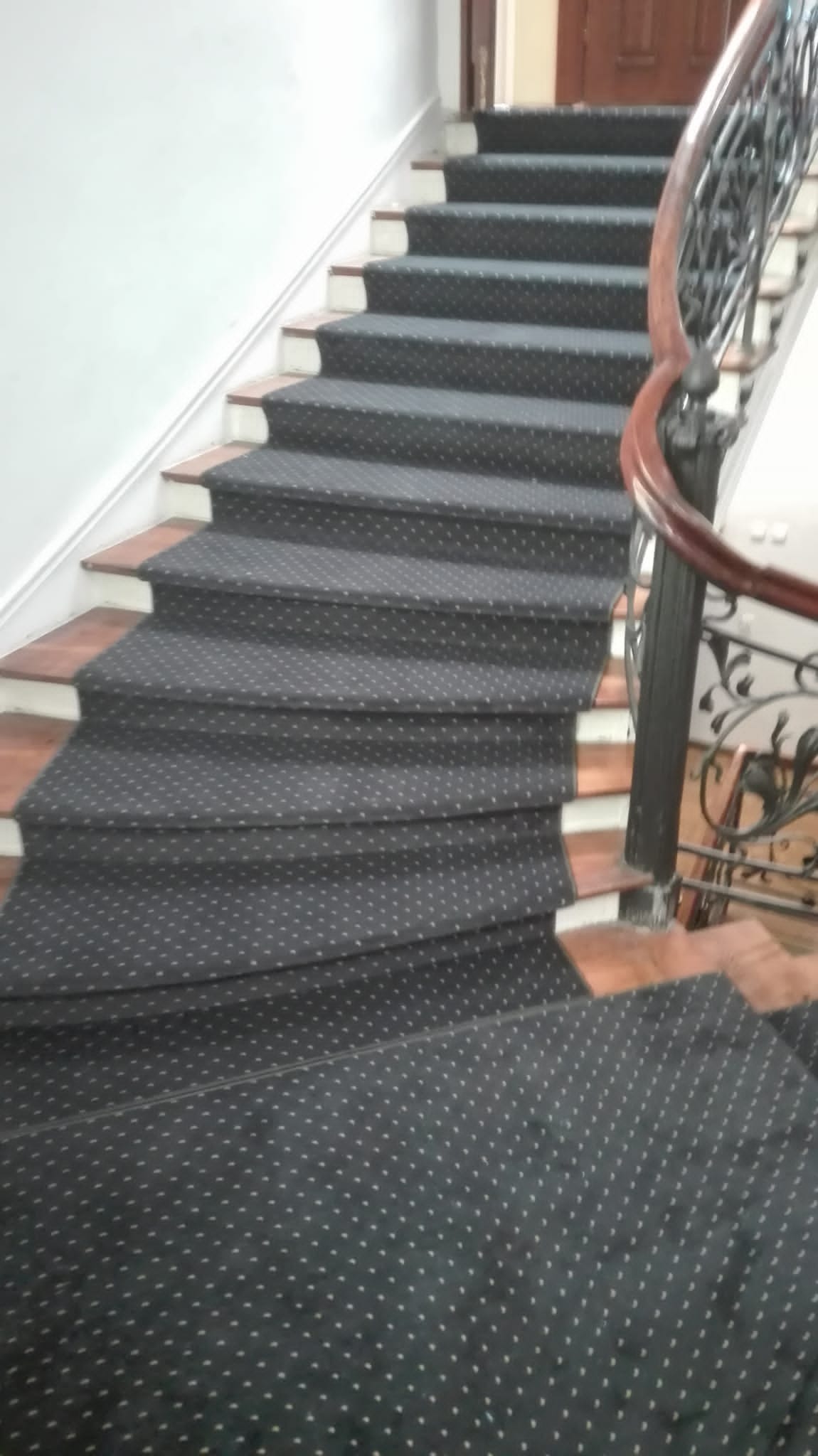 Schody wyłożone ciemną wykładziną w drobne białe kropki, z ozdobną, kutą balustradą.