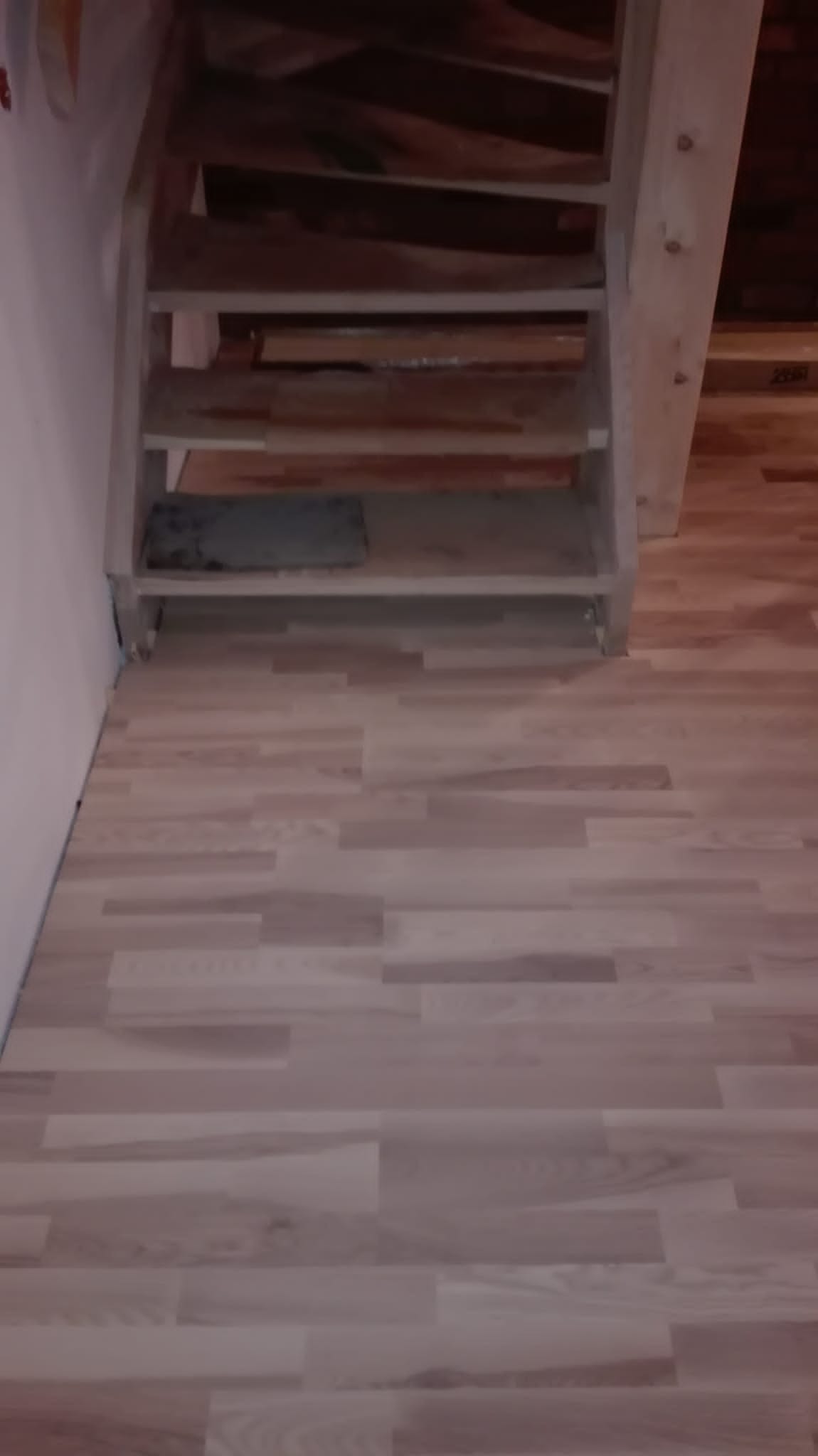 Jasne panele podłogowe ułożone przy drewnianych schodach bez poręczy, widoczne niedoskonałości wykończenia przy schodach.