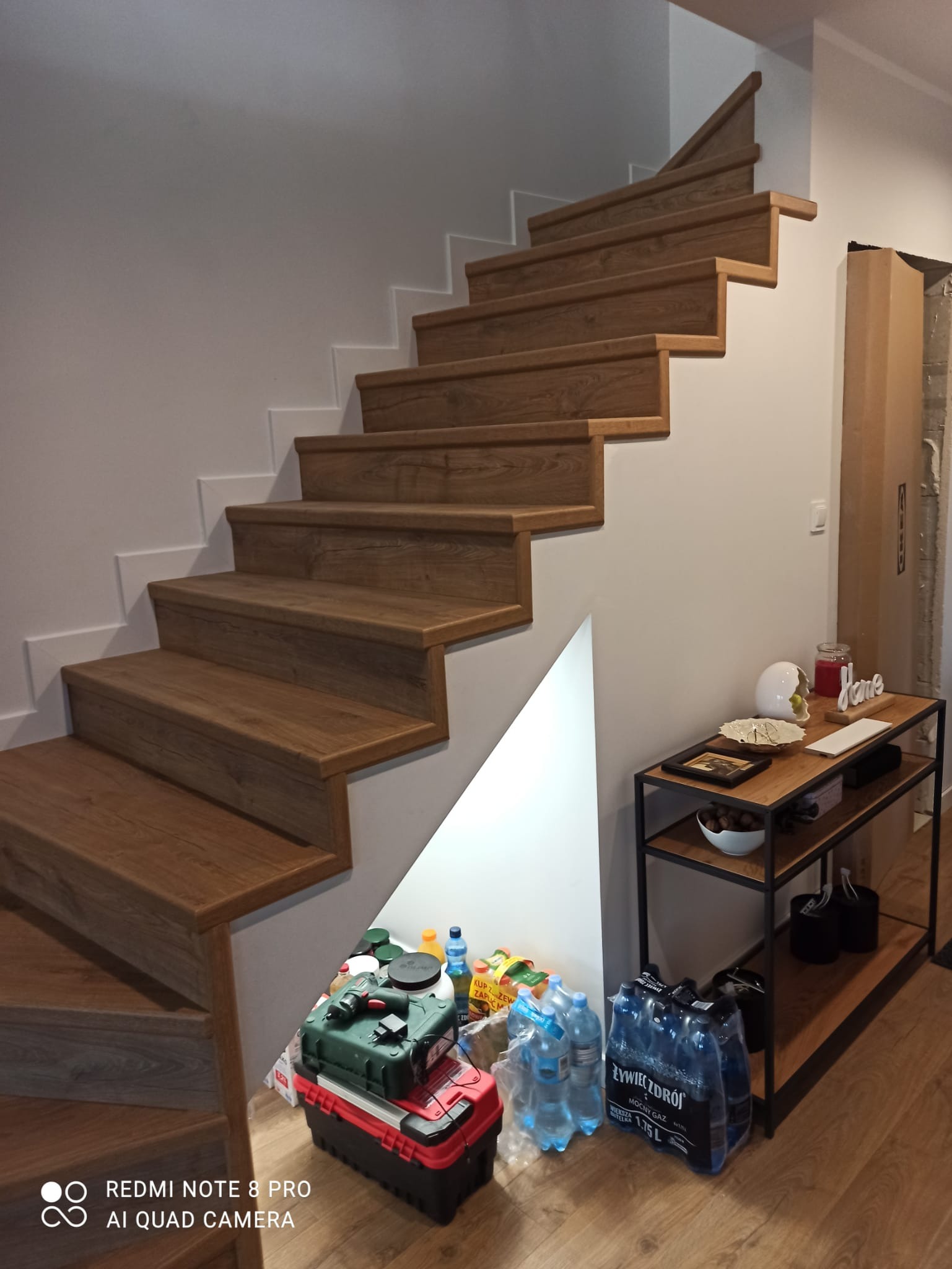 Schody drewniane z widocznym wykończeniem stopni, ujęte w trakcie prac remontowych, z narzędziami i materiałami budowlanymi zgromadzonymi u podnóża schodów, obok regału z dekoracjami.