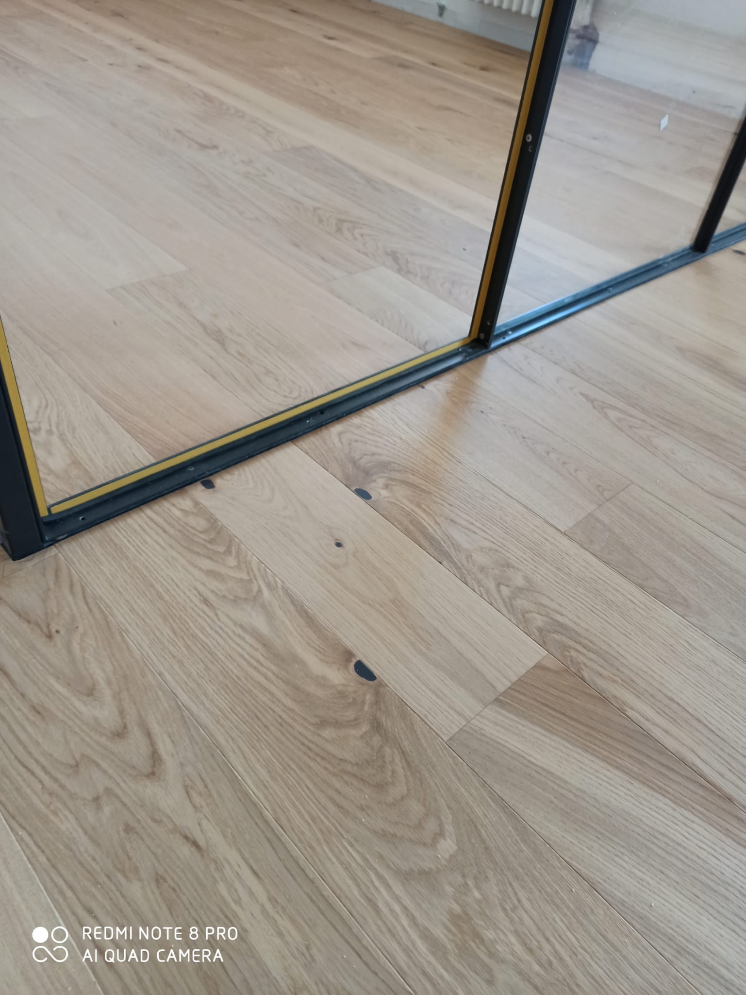 Jasne panele podłogowe z widocznymi słojami drewna, przy ścianie zamontowana czarna rama drzwi przesuwnych z żółtą uszczelką.