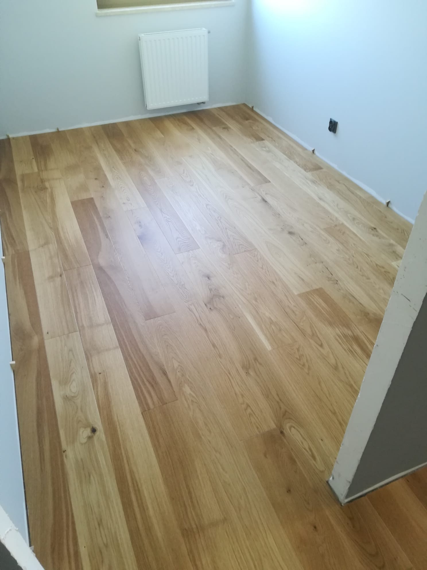 Nowo ułożona podłoga z drewnianych paneli w jasnym odcieniu dębu, widoczne listwy dystansowe przy ścianach, kaloryfer na ścianie w tle.