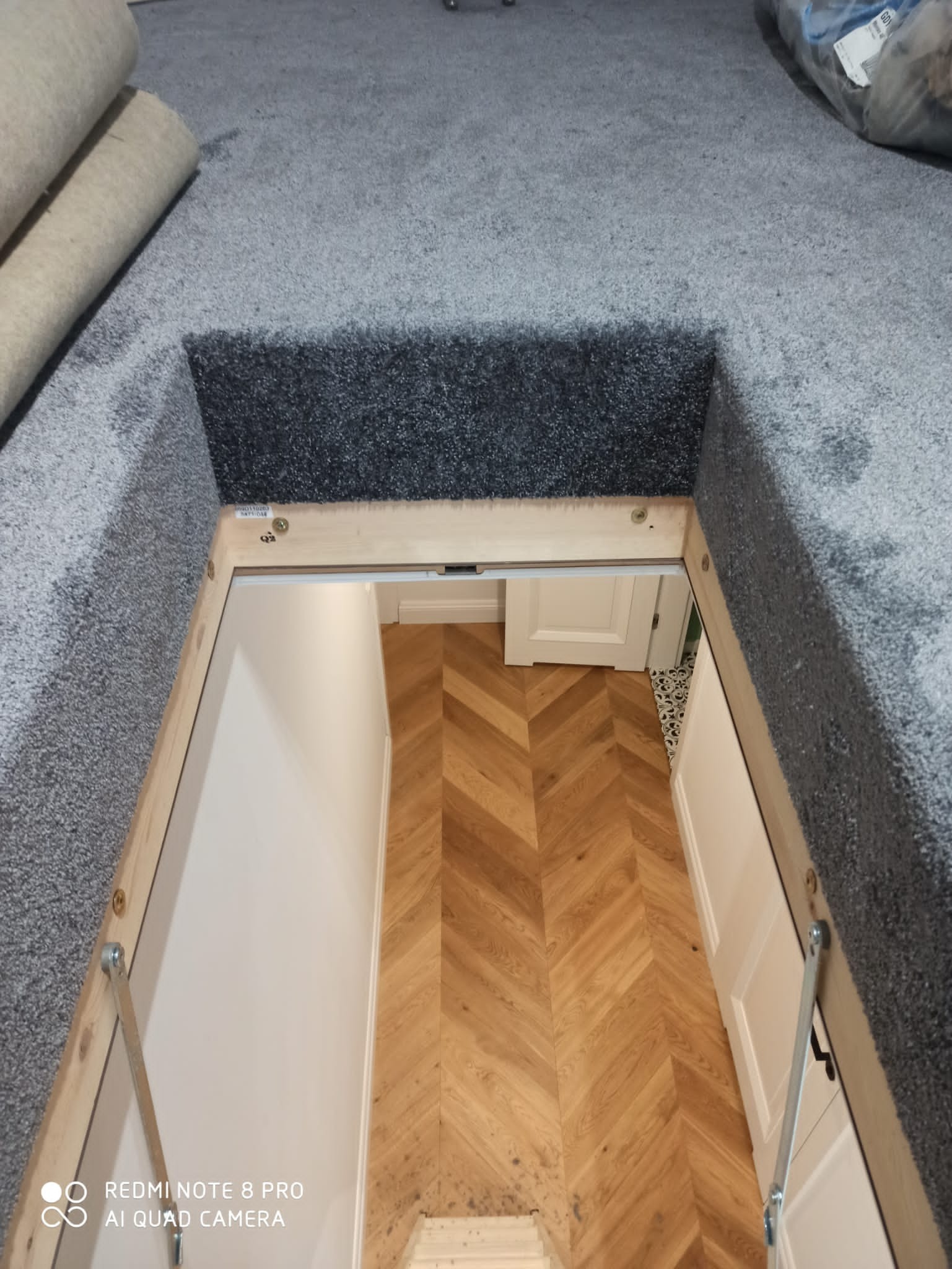 Gdańsk: Widok z góry na otwarty właz do piwnicy z zamontowaną szarą wykładziną dywanową, ukazujący parkiet ułożony w jodełkę francuską i białe drzwi.