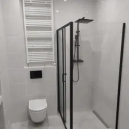 Nowoczesna łazienka z kabiną prysznicową typu walk-in z czarnymi profilami, wiszącą toaletą i białym grzejnikiem drabinkowym na tle białych płytek.