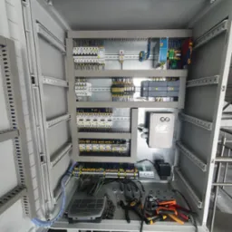 Otwarta szafa sterownicza z zamontowanymi modułami, przewodami i narzędziami, widok z góry.