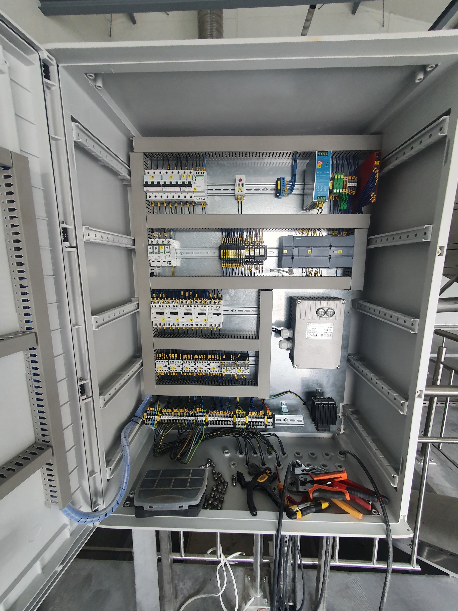 Otwarta szafa sterownicza z zamontowanymi modułami, przewodami i narzędziami, widok z góry.