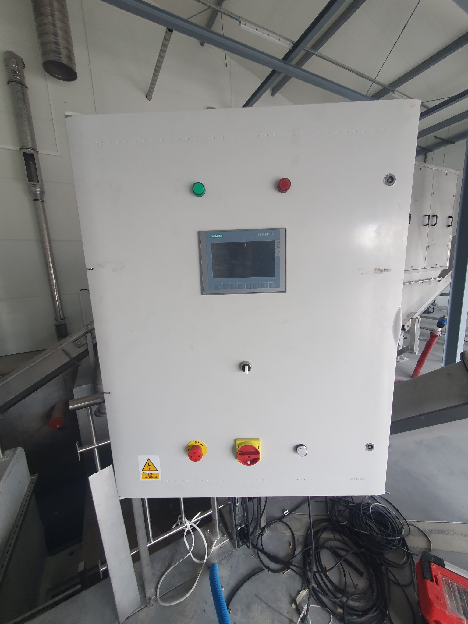 Biała szafa sterownicza Siemens Simatic HMI z przyciskami i ekranem dotykowym, widoczne kable i rury instalacyjne w tle, w hali przemysłowej.