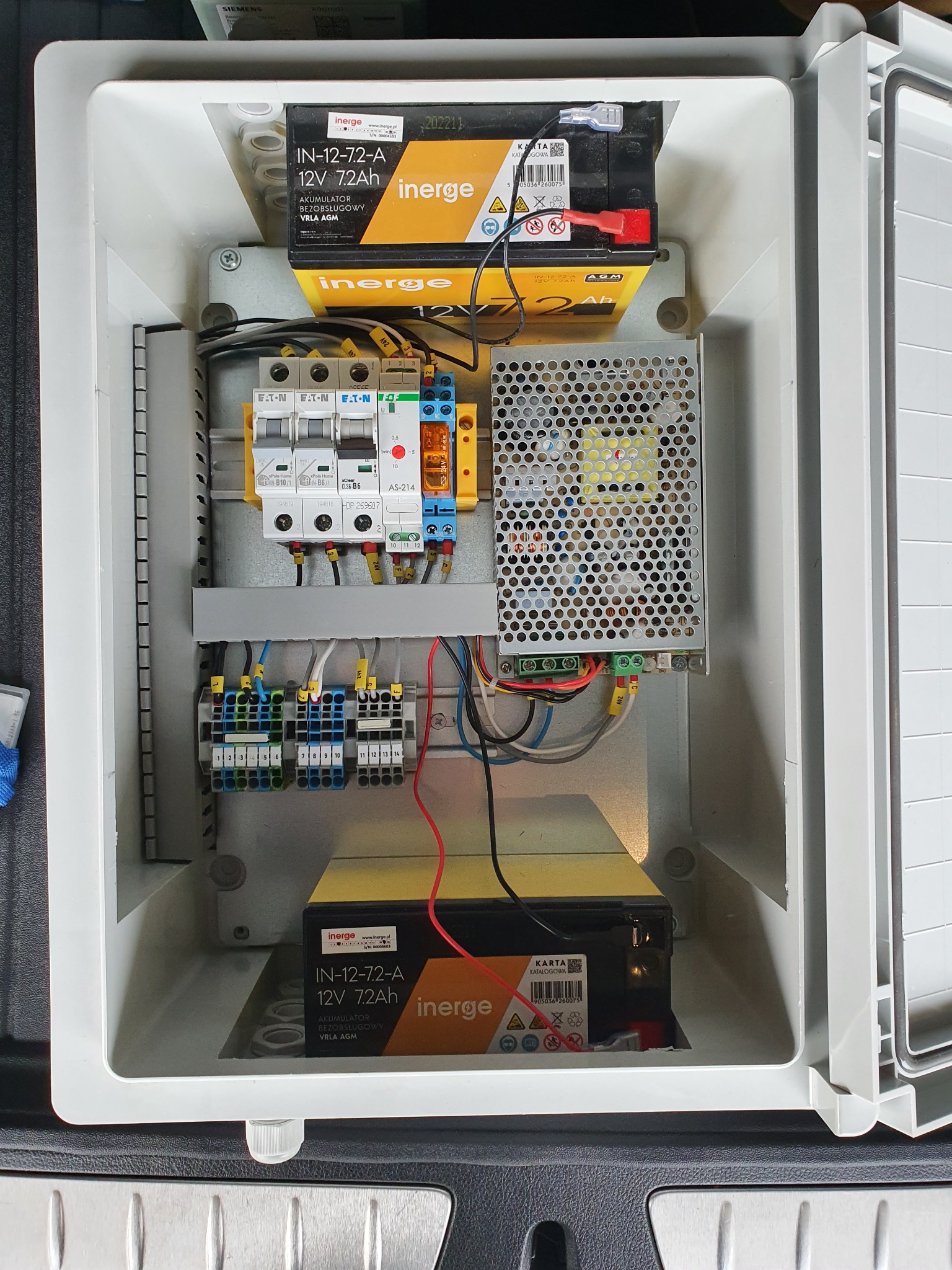 Wnętrze białej skrzynki elektrycznej z dwoma akumulatorami Inerge 12V 7.2Ah, bezpiecznikami Eaton, zasilaczem impulsowym i listwami zaciskowymi, widoczne okablowanie.