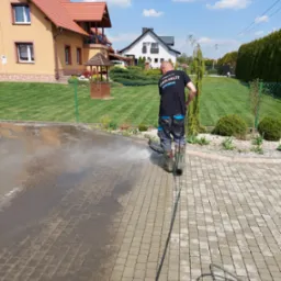 Pracownik w kaloszach czyści kostkę brukową na podjeździe domu za pomocą myjki ciśnieniowej; widoczne plamy brudu i strumień wody.