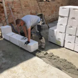 Murarz układa bloczki z betonu kom&oacute;rkowego na fundamencie, widoczny sznurek traserski i zapas bloczk&oacute;w, ściana z czerwonej cegły w tle.
