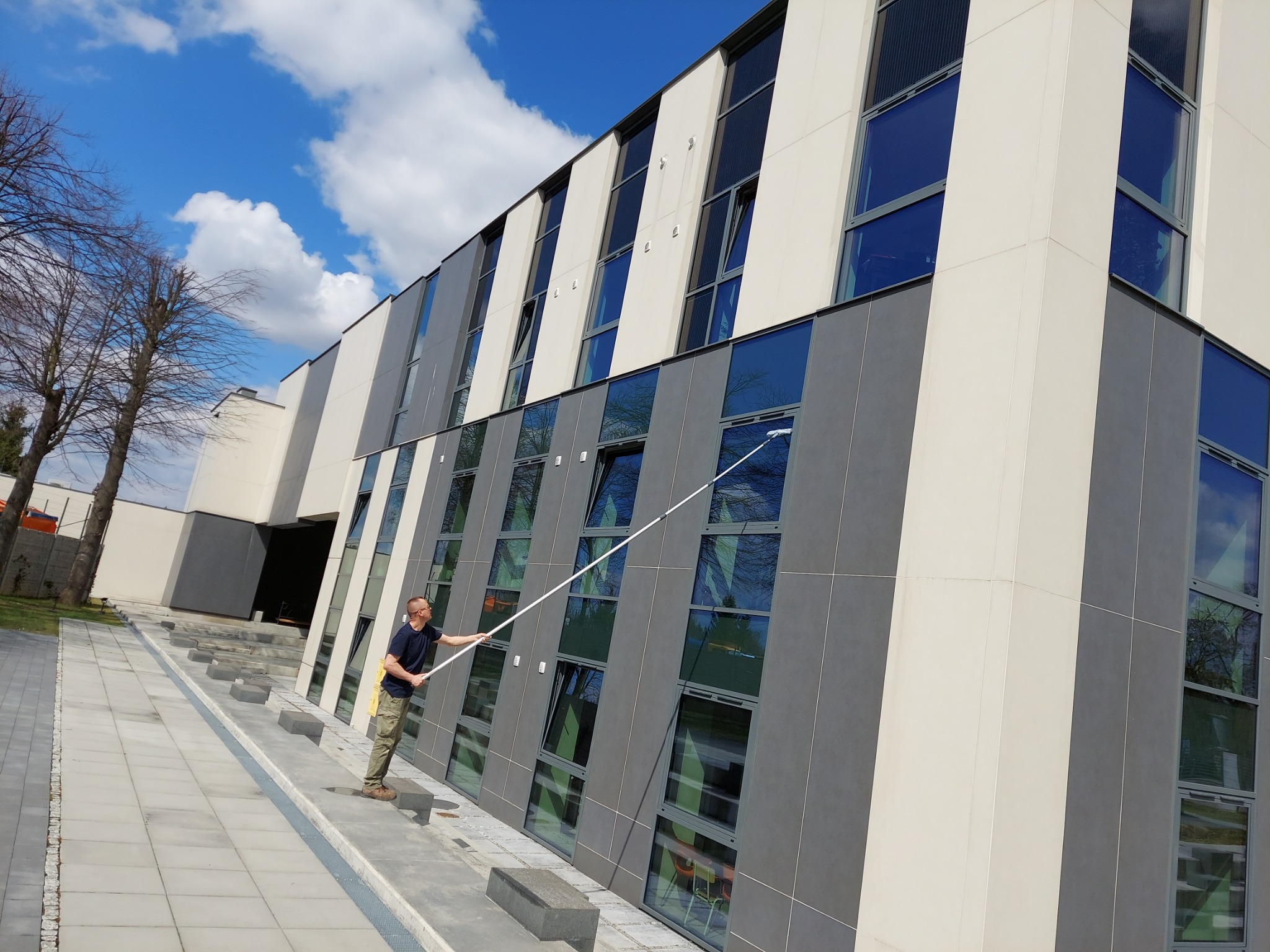 Mężczyzna w koszulce i spodniach moro myje okna na wysokości w nowoczesnym biurowcu za pomocą długiej lancy i myjki do okien. Część okien jest uchylona.