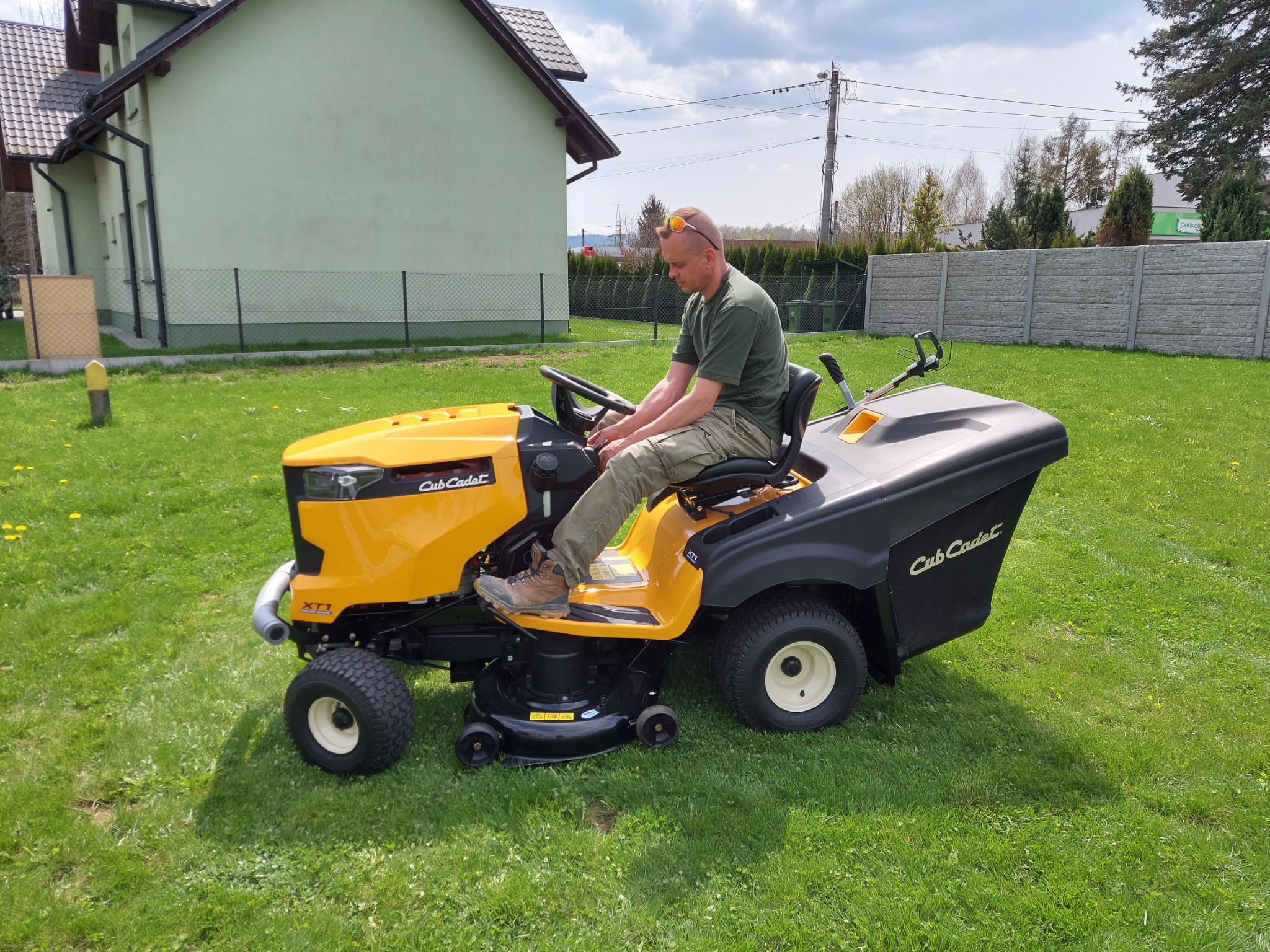 Operator na żółto-czarnej kosiarce trawnikowej Cub Cadet XT1, na tle zielonego trawnika i domu z jasną elewacją, w trakcie pracy.