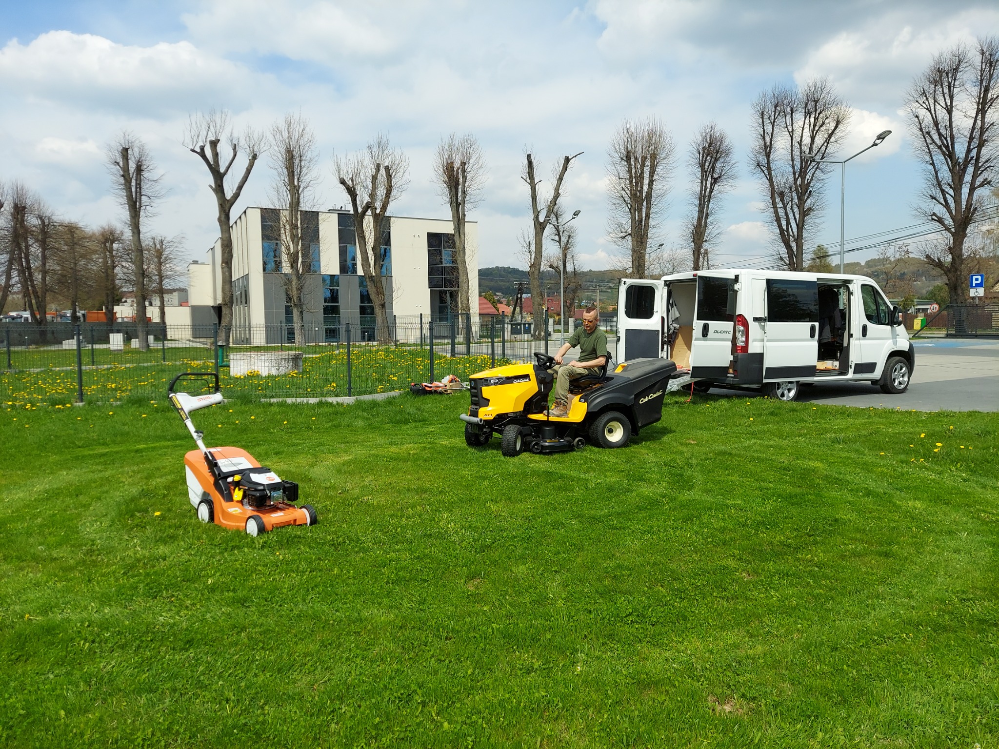 Mężczyzna na żółtej kosiarce samojezdnej Cub Cadet kosi trawnik, obok stoi pomarańczowa kosiarka Stihl, a w tle biały bus z otwartymi drzwiami i budynek.