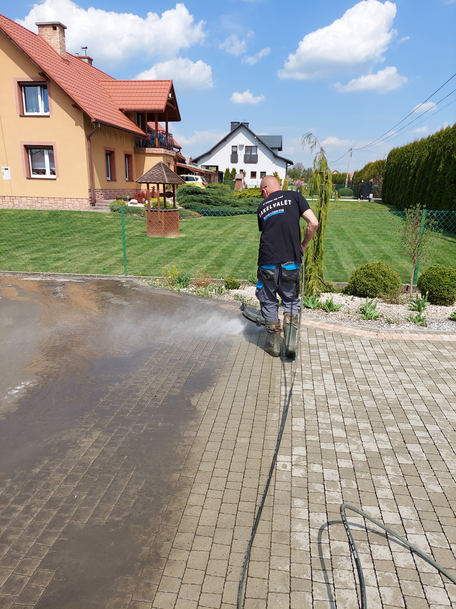 Pracownik w kaloszach czyści kostkę brukową na podjeździe domu za pomocą myjki ciśnieniowej; widoczne plamy brudu i strumień wody.
