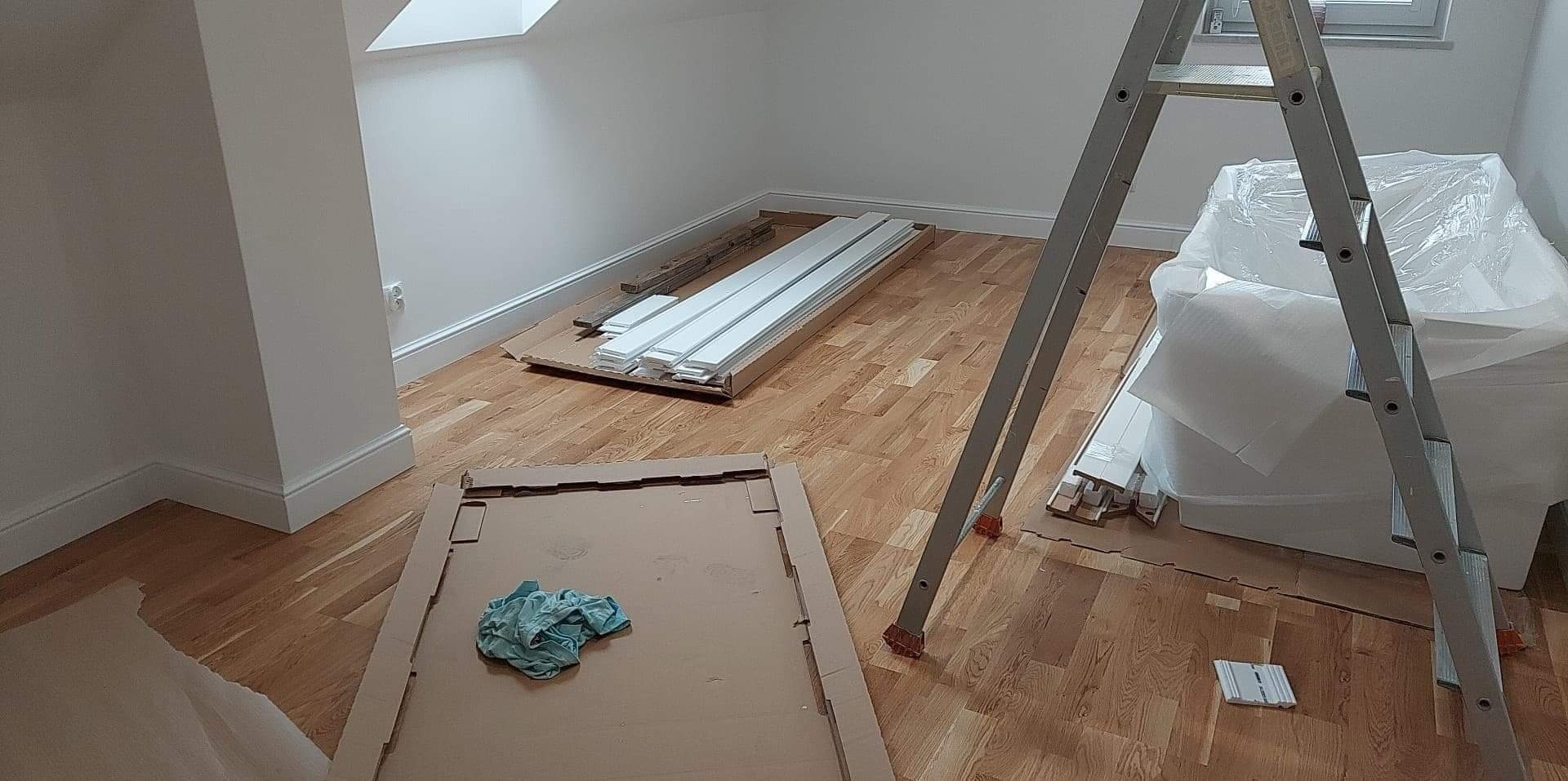 Pokój na poddaszu w trakcie remontu: drewniana podłoga, białe listwy przypodłogowe w kartonach, drabina oparta o ścianę, meble zabezpieczone folią.
